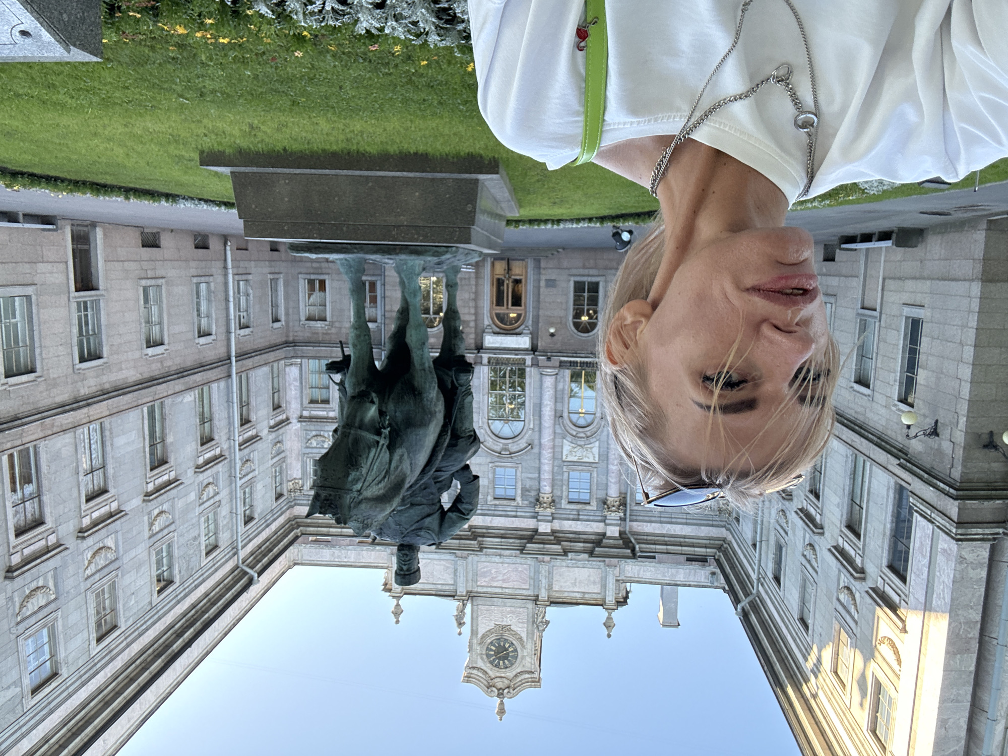 A blonde woman is taking a selfie in front of a large, classical building in Saint Petersburg, Russia. The building is made of light-colored stone and features numerous windows, arched doorways, and decorative elements. In the center of the frame, a large bronze equestrian statue of a man in military attire stands on a pedestal in front of the building. The woman is wearing a white t-shirt and silver necklaces, and her sunglasses are perched on her head. She is looking towards the camera with a thoughtful expression. The scene is set on a sunny day with a clear blue sky. The area around the statue is a manicured lawn with flowerbeds. The overall impression is one of historical grandeur and a personal moment captured in time.