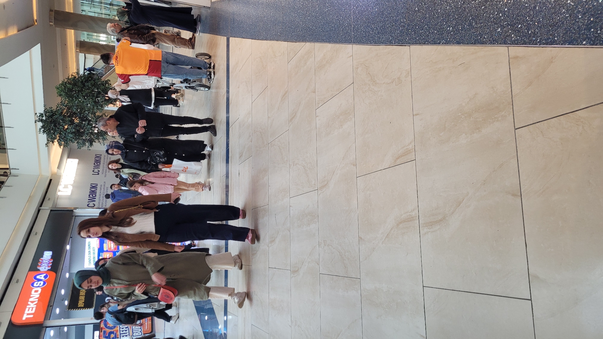 Here is a detailed description of the image:
The image shows the interior of a shopping mall in Başakşehir, Turkey. A line of people are standing, with shops visible on one side. There are at least ten people in the image, including individuals of varying ages and attire. Some are dressed in casual wear like jeans and t-shirts, while others wear more traditional or modest clothing such as headscarves and long coats. In the foreground, there are advertisements for shops, one of which is "TEKNOSA," followed by texts in Turkish, indicating shopping and sales promotions. Another is a shop called "LC WAIKIKI." The flooring of the mall is made of large, beige tiles, and the ceiling is visible with artificial lighting fixtures. The overall impression is that it is a busy indoor shopping environment.