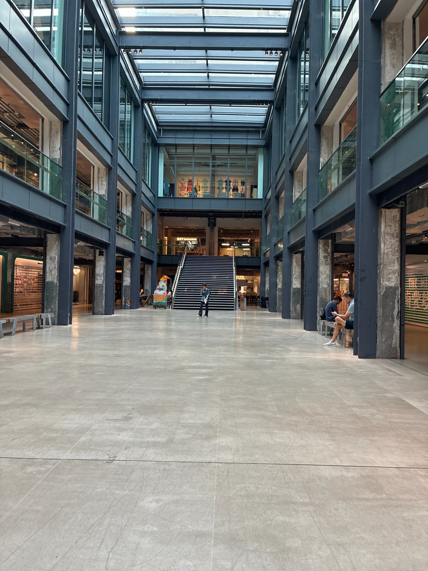 The image depicts the central atrium of a multi-story indoor public space, likely a shopping mall or commercial complex. The architecture features exposed industrial elements such as dark metal beams and columns, juxtaposed with concrete structures and large expanses of glass, suggesting a modern design that incorporates raw, industrial aesthetics. The atrium is open to a glass ceiling, allowing natural light to illuminate the space.
In the foreground, a vast, polished concrete floor stretches towards a central staircase. Two individuals are seated on benches on the right side of the image, one looking at their phone. A person is walking towards the stairs in the middle ground. To the left, benches are arranged near storefronts. Further back, a vibrant, cartoonish sculpture of a bird-like creature stands near the base of the stairs. Shops with glass facades are visible on multiple levels surrounding the atrium. The overall impression is that of a contemporary urban environment designed for public gathering and commerce. The architectural style and the presence of similar indoor retail spaces with exposed industrial design are characteristic of revitalized industrial areas in Hong Kong, such as PMQ or The Mills.