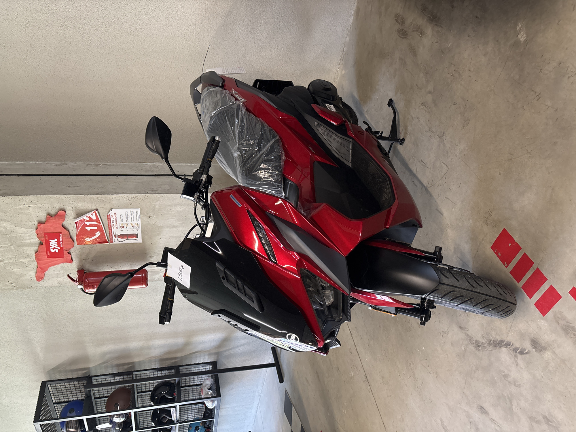 The image depicts a red scooter parked indoors, possibly in a showroom or garage. The scooter is the primary subject, with its front wheel and fairings prominently displayed. It has a sleek, modern design with black accents and a clear plastic seat cover, suggesting it is new or recently unwrapped. A price tag displaying "6500" is attached to the windshield.
In the background, a metal shelving unit holds several helmets of various colors, including blue, brown, and black. To the right of the scooter, a red fire extinguisher is mounted on the wall, accompanied by two signs: one shaped like Taiwan with the logo "SYM FROM TAIWAN", and another with the number "112" and instructions on how to use a fire extinguisher. The walls appear to be concrete or a similar textured material, and the floor is a concrete surface with some scuff marks and red rectangular markings. The overall environment suggests a commercial or industrial setting. The location in the city of Tbilisi, Georgia, is not visually indicated in the image.