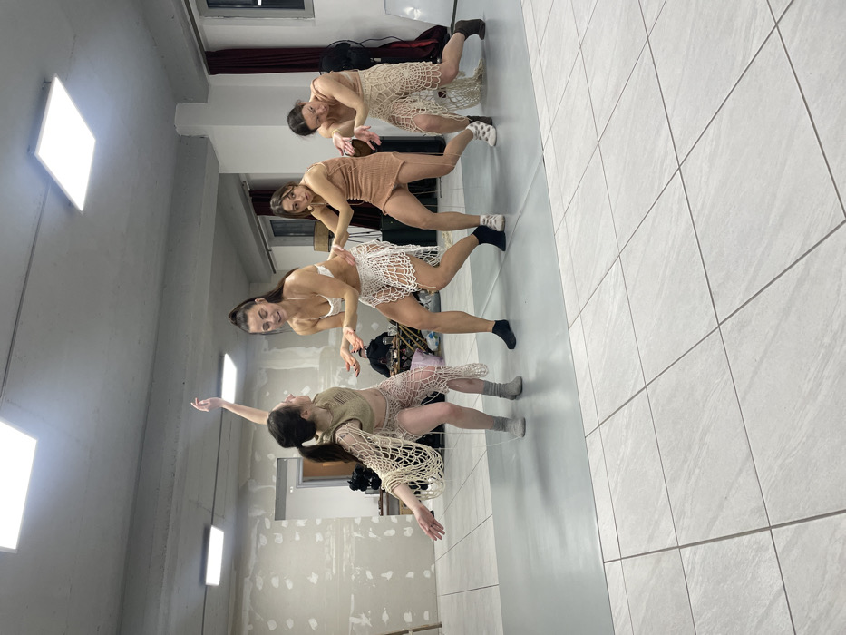 The scene takes place in an indoor studio or rehearsal space in Budva, Montenegro. Four adult females are depicted in a choreographed pose or movement sequence across the mid-ground. The room features light-colored walls with visible patching, a light gray tiled floor, and a large mirror reflecting the scene, positioned on the left side. Two square LED panel lights are affixed to the ceiling. A dark gray rectangular mat is present on the floor beneath the subjects. A window with dark red curtains is located on the right.
The females are dressed in similar light-toned, open-weave, knitted or crocheted two-piece outfits.
The leftmost female, oriented towards the right, has her left arm raised vertically and her right arm extended outwards. She wears a light beige outfit and light gray socks.
The second female from the left, facing slightly right, has a subtle smile and both hands extended forward with palms open. She wears a white bra-top, a white crochet skirt, and black socks. Her dark hair is styled in a high ponytail.
The third female from the left, facing slightly right with her mouth slightly open, has her right hand extended and her left hand grasping the arm/sleeve of the female to her right. She wears a light brown knitted top and matching skirt, with white socks.
The rightmost female is in a low, crouching position, facing slightly left. Her left hand is open, and her right arm is held by the adjacent female. She wears a light beige top and skirt, along with dark shoes.
In the background, a small table holding a cup and other indistinct objects is visible, alongside dark fabric and a piece of dark equipment near the window.
