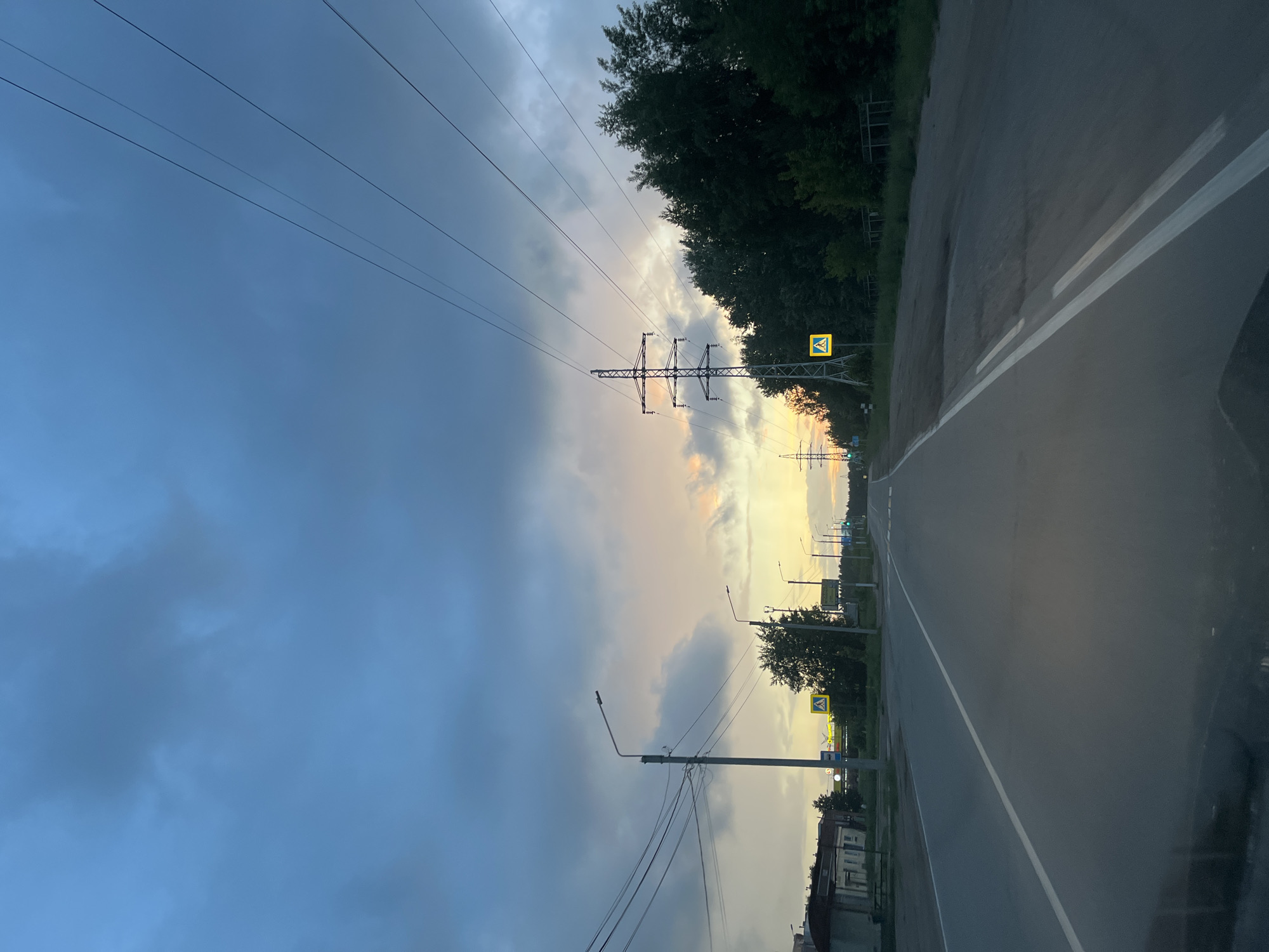 The image depicts an outdoor scene at either dusk or dawn in Chaikovsky, Russia. A paved road stretches from the foreground into the distance, bordered by trees and utility infrastructure.
The sky dominates the upper portion of the frame, showcasing a striking contrast. On the left, dark blue-grey clouds are prevalent, indicating an overcast or transitioning sky. Towards the right, the sky lightens considerably, revealing warm hues of orange and yellow, characteristic of a sunrise or sunset. Several power lines cut across the sky, leading to and from utility poles and a large metal electricity pylon, which stands prominently in the middle-ground, silhouetted against the lighter sky.
Along the right side of the road, a dense line of green trees forms a natural boundary. A yellow square sign with a black pedestrian symbol is clearly visible on the right, indicating a pedestrian crossing. Further along the road, more streetlights and smaller utility poles are present. The road itself features distinct white markings: a solid line separating the main lane from the shoulder, and further right, a double dashed line.
The scene is devoid of people or vehicles, conveying a sense of stillness and quiet. The natural lighting suggests a peaceful transition between day and night, or vice versa, in a semi-urban or rural setting. No discernible text is visible on any signs or objects.