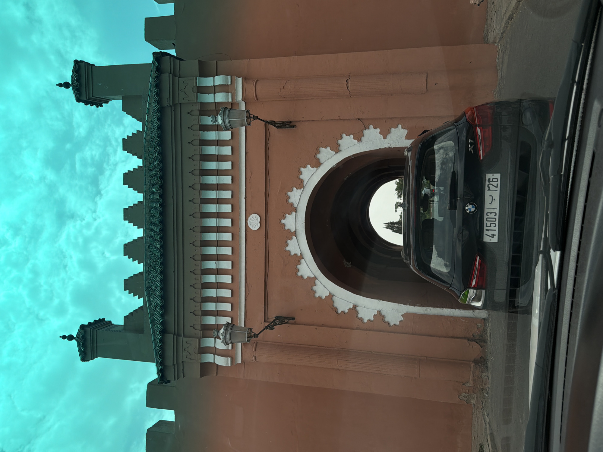 The image shows the exterior of a historic gateway in Mechouar Kasbah, Morocco. The gateway is constructed of reddish-orange brick and features ornate white decorative elements above an arched passageway. Two vintage-style lampposts are mounted on either side of the archway. Atop the gateway, a dark green crenellated roofline is visible, with two decorative turrets at the ends. The sky above is a vibrant turquoise with scattered clouds. A black BMW X1 SUV, with license plate "41503 ب 26", is positioned directly in front of the archway, partially obscuring the entrance. The view appears to be from inside another vehicle, as the windshield wipers and dashboard are visible in the foreground. The overall scene is static, with no discernible human activity beyond the presence of the car.