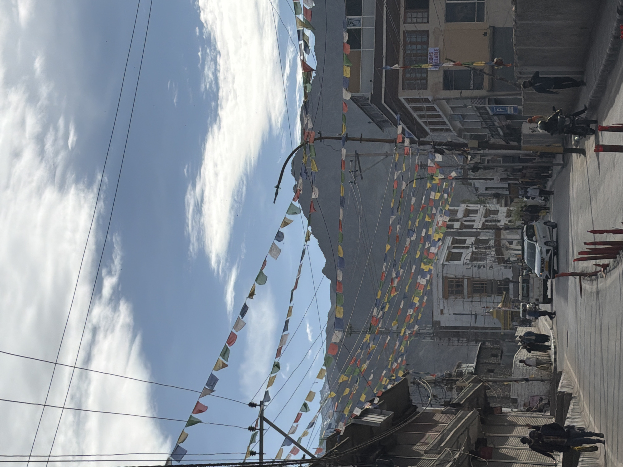 The image depicts a street scene in Leh, India, characterized by multi-story buildings on either side of the road, with a backdrop of majestic mountains. Prayer flags, in various colors like blue, white, red, green, and yellow, are strung across the street, creating a vibrant and festive atmosphere. Electrical wires and poles are visible, interwoven with the flags.
The street appears to be a main thoroughfare, with vehicular traffic including a white SUV and a white van. Several people are seen walking on the sidewalks and crossing the street. One person is riding a motorcycle. The buildings showcase a mix of architectural styles, with some featuring balconies and traditional window designs. One building has a sign that reads "ROOMS AVAILABLE." The sky is partly cloudy, with the sun casting shadows on the street. The overall impression is of a bustling urban environment set against a stunning natural landscape.