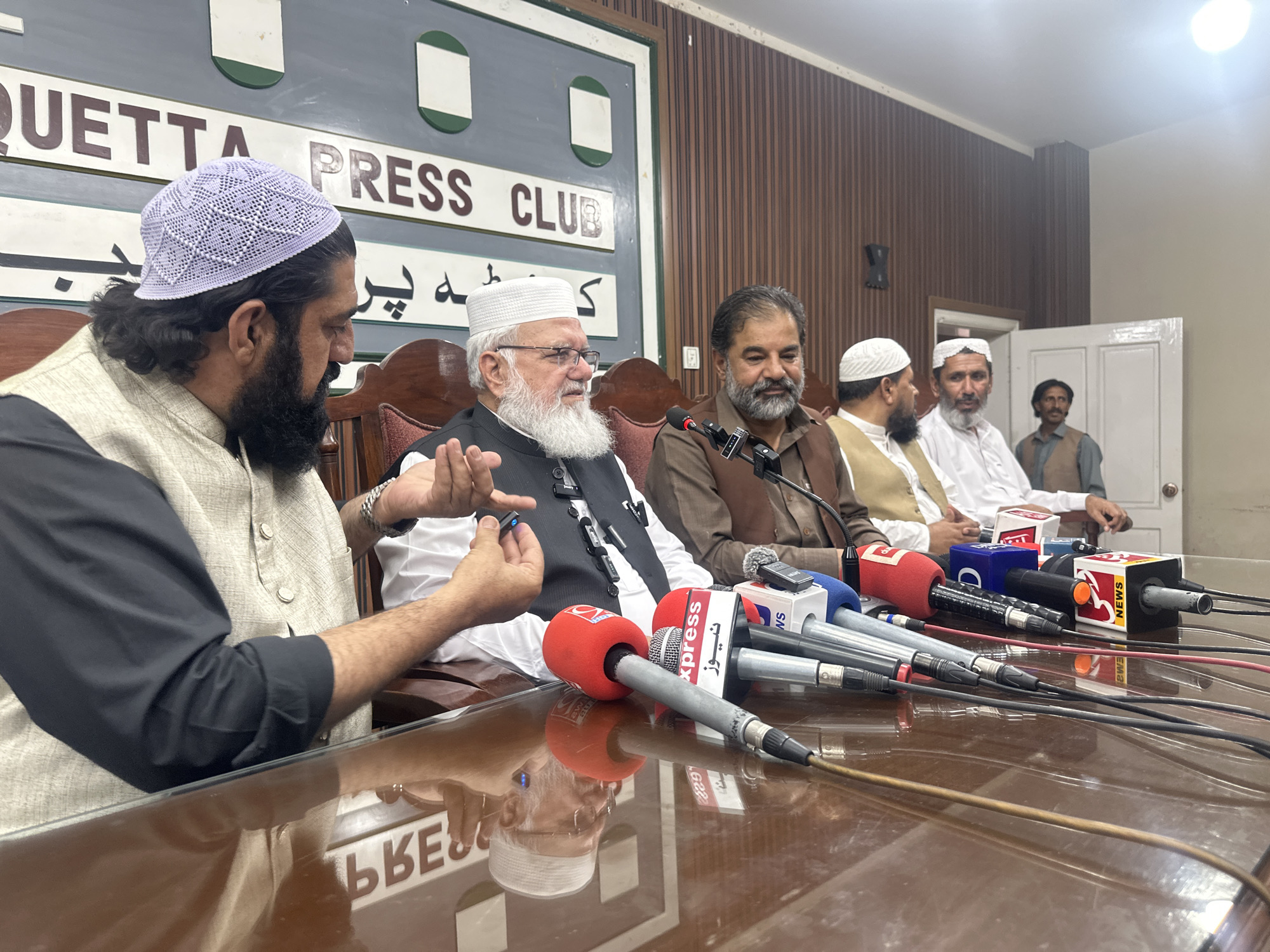 The image shows a press conference being held in Quetta City Tehsil, Pakistan. Several men are seated at a table with microphones in front of them. The background features a sign that reads "QUETTA PRESS CLUB". The men are dressed in traditional Pakistani attire. The man in the foreground on the left is wearing a light-colored vest over a black shirt and a white patterned cap. He has a full beard and is gesturing with his right hand. To his right, an older man with a white beard and glasses is also seated and appears to be speaking. Several microphones, including some with "Express News" branding, are positioned on the table. The room has wooden paneled walls.