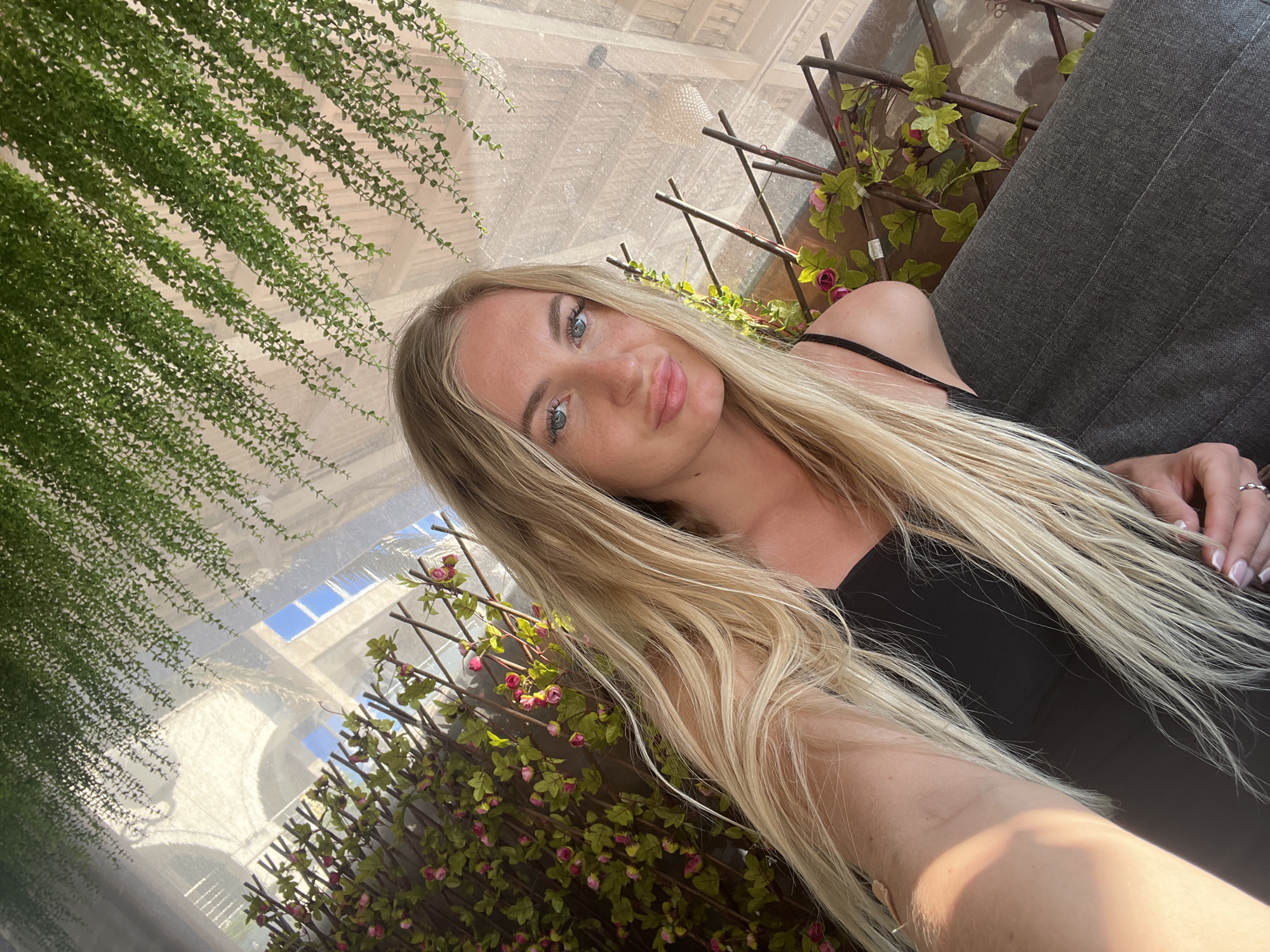 A selfie of a fair-skinned woman with long blonde hair, blue eyes, and slightly pursed lips. She is wearing a black tank top. To the left, there is hanging greenery. Behind and to the right, there are decorative branches with green leaves and small pink flowers. In the background is a view of a white building under a translucent roof. This image was taken in the city of Rafailovići, Montenegro. There is also a grey surface on the right side, possibly a sofa.