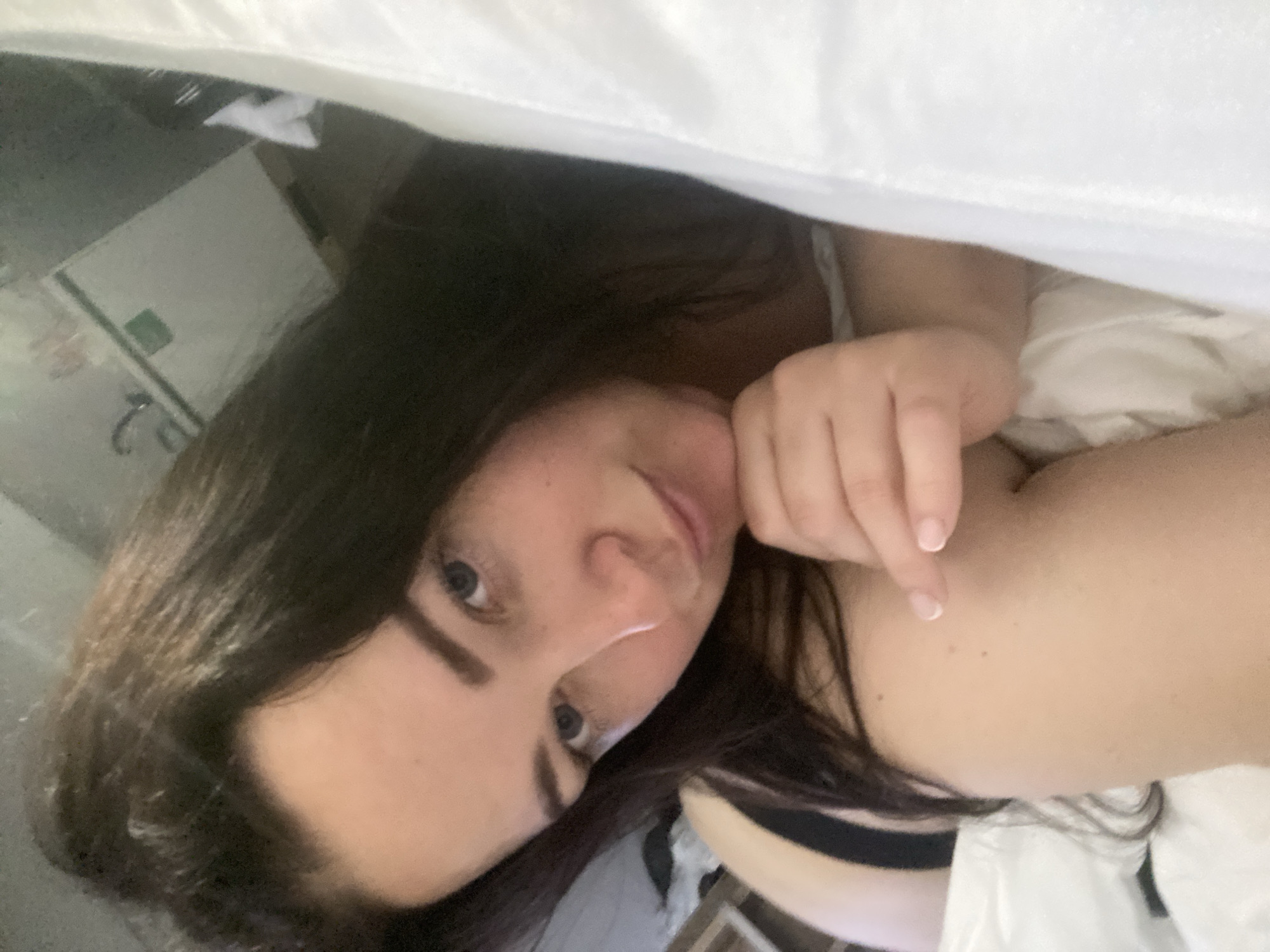A woman lies in bed under a white sheet. She has long, dark hair and fair skin. Her left hand is raised to her face with her index finger extended. Behind her is a light-colored wall and a white dresser. The location is unknown.