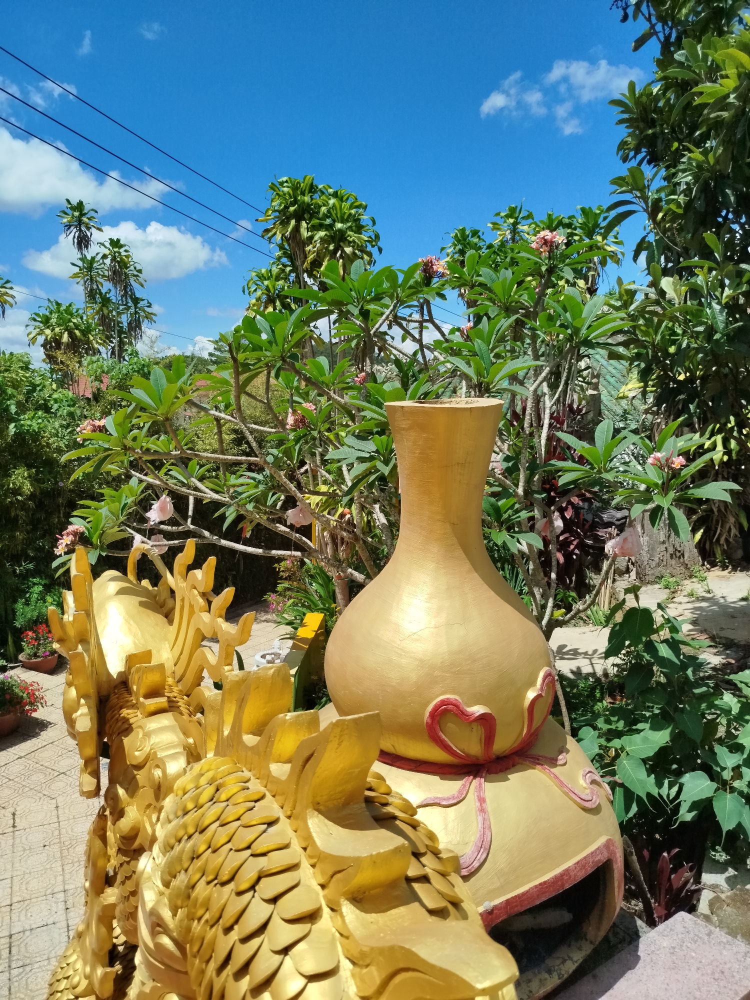 Captured in the city of Da Lat, Vietnam, a gilded ornamental vase stands prominently in a garden setting, adorned with intricate red heart designs on its lower bulbous section. A golden dragon structure, detailed with overlapping scales and ornate crests, partially frames the vase in the foreground. The surrounding flora includes flowering plants with pink blooms, lush green foliage, and several slender trees visible against a partly cloudy blue sky. Power lines traverse the upper portion of the frame, suggesting an urbanized setting. The overall scene conveys an aesthetic of cultural artistry integrated with natural landscaping.