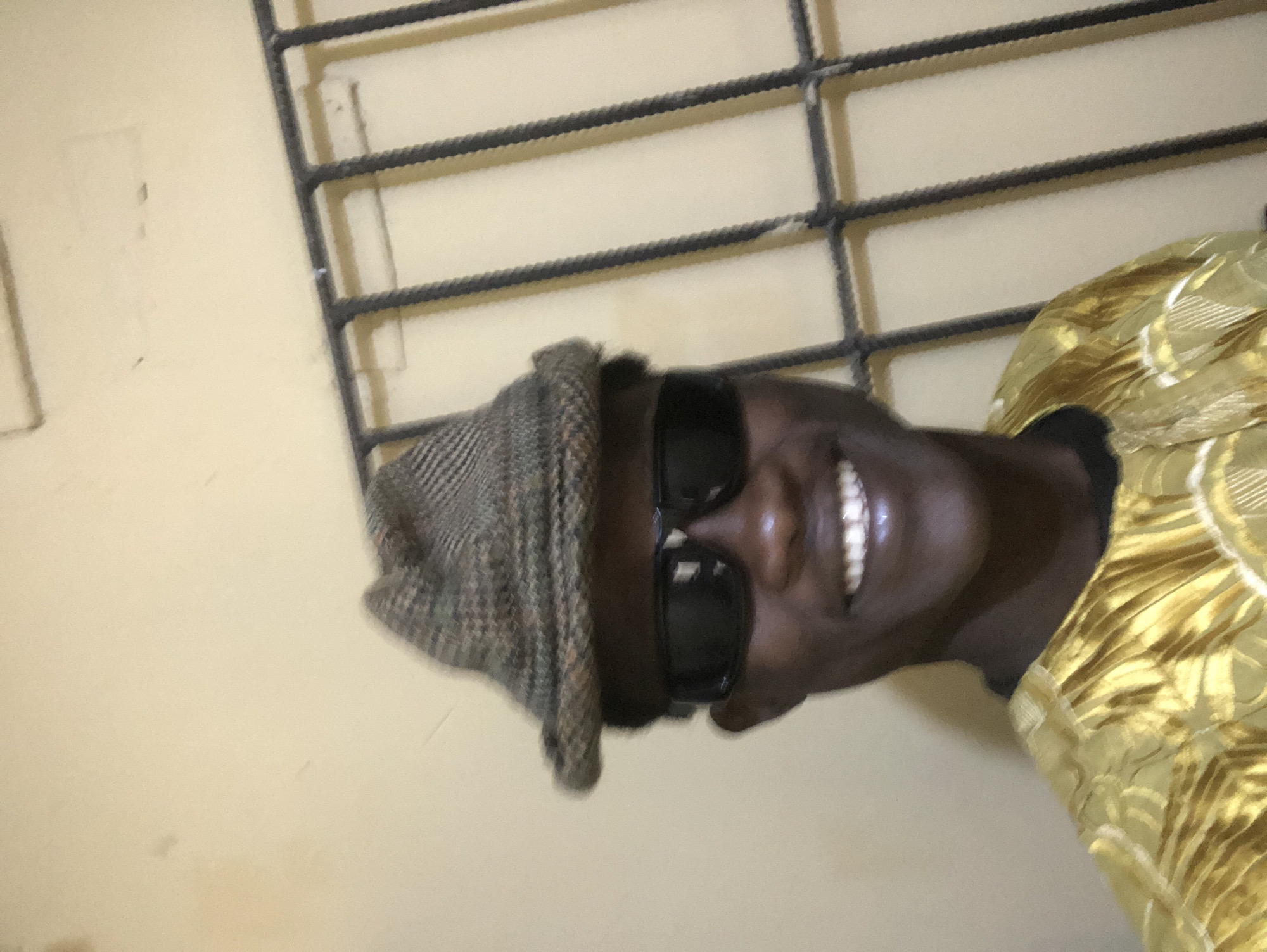 A young Black male, wearing a brown checkered hat and black sunglasses, smiles at the camera. He is dressed in a bright gold, patterned garment, possibly traditional attire. Behind him, a light-colored wall is visible, with a dark metal grate or bars crossing the frame. The lighting is even, and the image appears to have been taken indoors, possibly in a home or residential setting in Ado, Nigeria, given the style of the grate and the wall texture, which is consistent with common construction in that region.