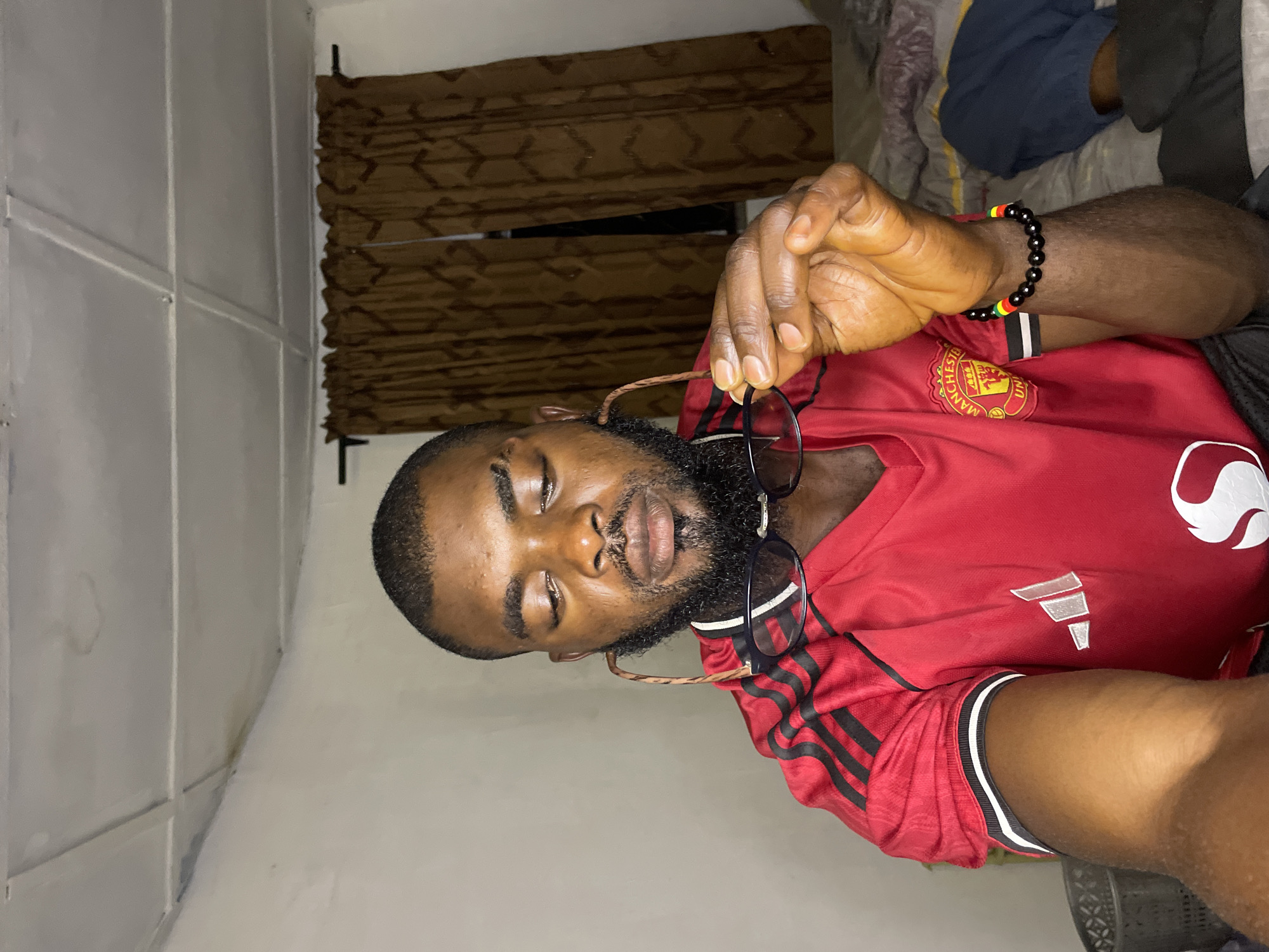 A medium shot shows a Black man with a beard and short hair looking down at a pair of glasses he holds in his right hand. He is wearing a red Manchester United jersey with black stripes on the shoulders and a white and black striped trim on the sleeves. A black beaded bracelet with red, yellow, and green beads adorns his left wrist. The man is in a dimly lit room with white walls. A brown patterned curtain hangs in the background, obscuring a window. The ceiling appears to be white and textured, with visible water stains in one corner. A bed with grey and blue bedding is visible to the right of the frame. A dark grey basket is partially visible on the left. The overall setting suggests an indoor, domestic environment.