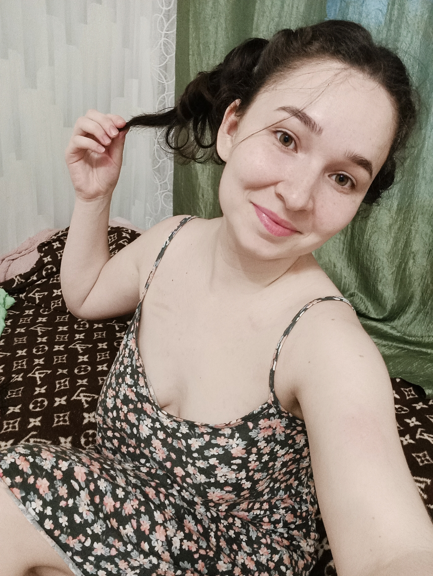A woman is sitting on a bed, possibly in a home in Izhevsk, Russia. She is fair-skinned with brown hair in pigtails and brown eyes, wearing a floral print dress with spaghetti straps. She is smiling and holding a strand of her hair. The bed has a brown patterned blanket. Behind her are white sheer curtains on the left and a green curtain on the right.
