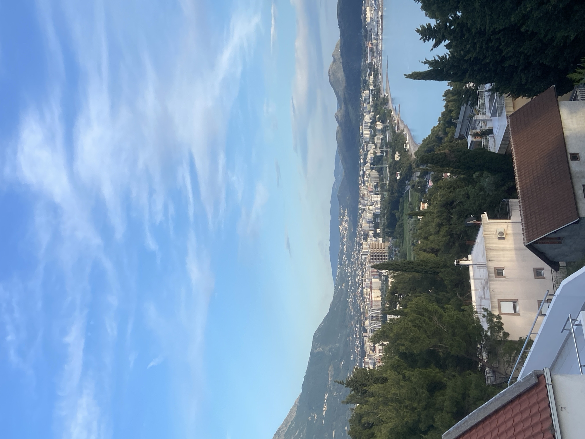 An oblique, outdoor daytime shot shows Šušanj, Montenegro, from an elevated vantage point. Mountains stretch across the scene, their slopes partially obscured by buildings and lush trees. The town is positioned along the coast, where the clear water extends to the horizon. The sky is a light blue hue, with streaks of white clouds scattered across the frame. Rooftops and the tops of trees are visible in the lower right corner, suggesting the photo was taken from a building or high ground. No people are visible.