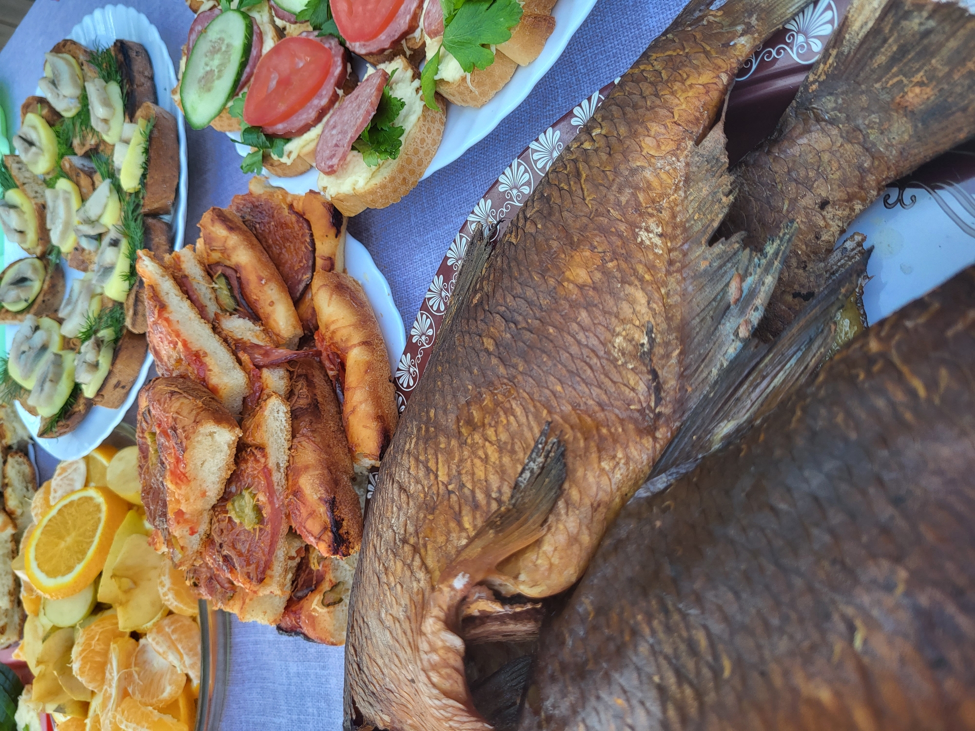 The image shows a close-up of a food spread, possibly outdoors. The main subject is two smoked fish placed on a white plate. To the left, there are several plates with various appetizers. These include sandwiches with toppings such as cucumbers, tomatoes, and what appears to be sausage. Another plate has grilled bread with mushrooms and dill, while a third one contains sandwiches with visible filling. A plate with sliced oranges and apples is also present. The setting appears to be a table covered with a grey tablecloth. The scene does not provide information regarding Зипуново, Russia.