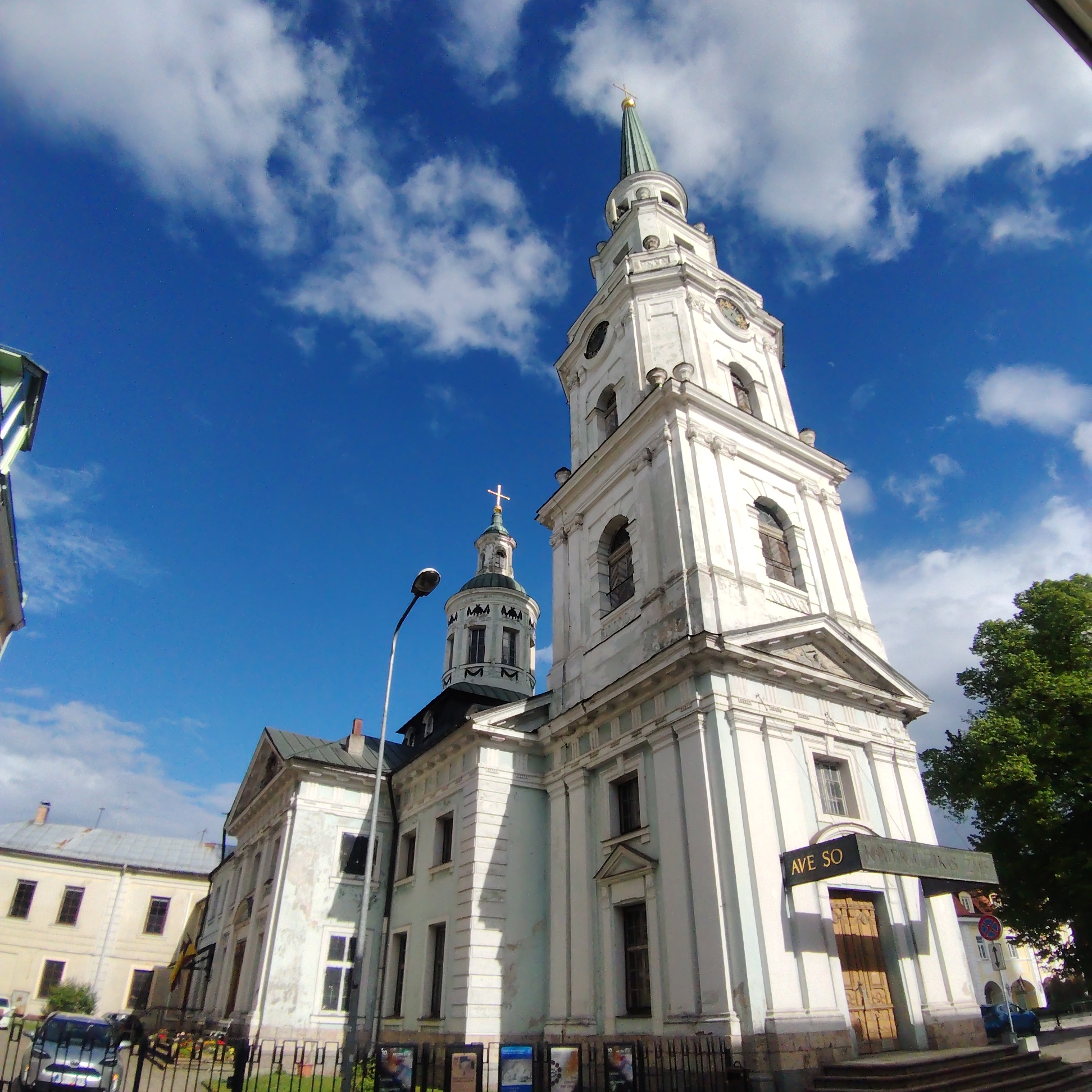 The image depicts St. Peter's Church, an Evangelical Lutheran church in Riga, Latvia. The church is predominantly white with a tall, multi-tiered tower topped with a green spire and a golden cross. A smaller, dome-like tower with a cross is positioned to the left of the main tower. The building's entrance features a sign with the text "AVE SO." The sky is bright blue with scattered white clouds. In the foreground, there are cars, street furniture, and a fence. A tree is visible on the right side of the frame.