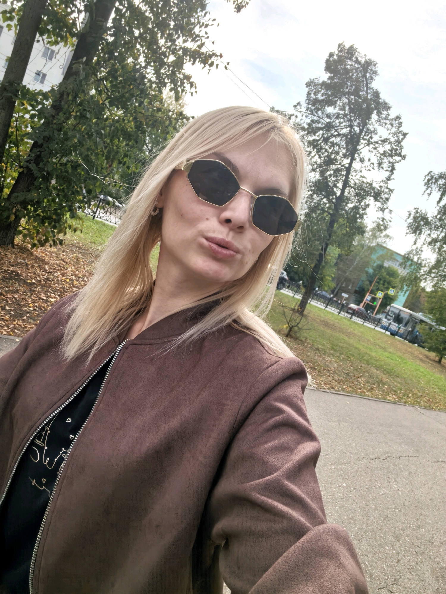 A woman with blonde hair and sunglasses poses for a selfie outdoors in Sterlitamak, Russia. She is wearing a brown jacket over a black shirt. The background shows trees, fallen leaves, a sidewalk, a street with cars and a bus, and a building. The sky is visible above.