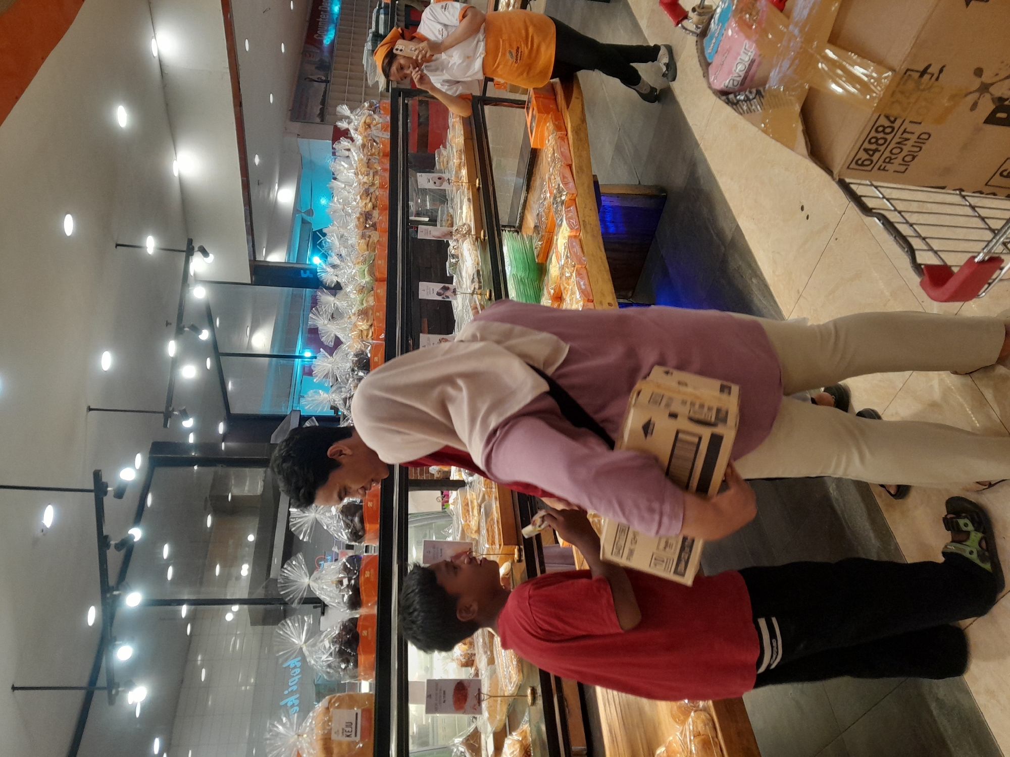 The image depicts the interior of a bakery, likely located in Banyuwangi, Indonesia, judging by the context provided. Three individuals are visible. In the foreground, a young boy wearing a red t-shirt and black pants with green sandals is facing a woman wearing a light purple hijab and light beige pants, who is holding a cardboard box. The woman is also interacting with another person out of frame to her left. Behind them, another person, a young man, is partially visible.
The bakery features display shelves filled with baked goods, many of which are individually packaged in clear plastic. The lighting appears to be from overhead fluorescent fixtures. A shopping cart is visible in the lower right corner of the frame, containing packaged items. A woman wearing an orange apron and a headscarf is standing further back on the right side of the image, engaged in a phone call. The overall environment suggests a retail setting focused on selling baked goods.