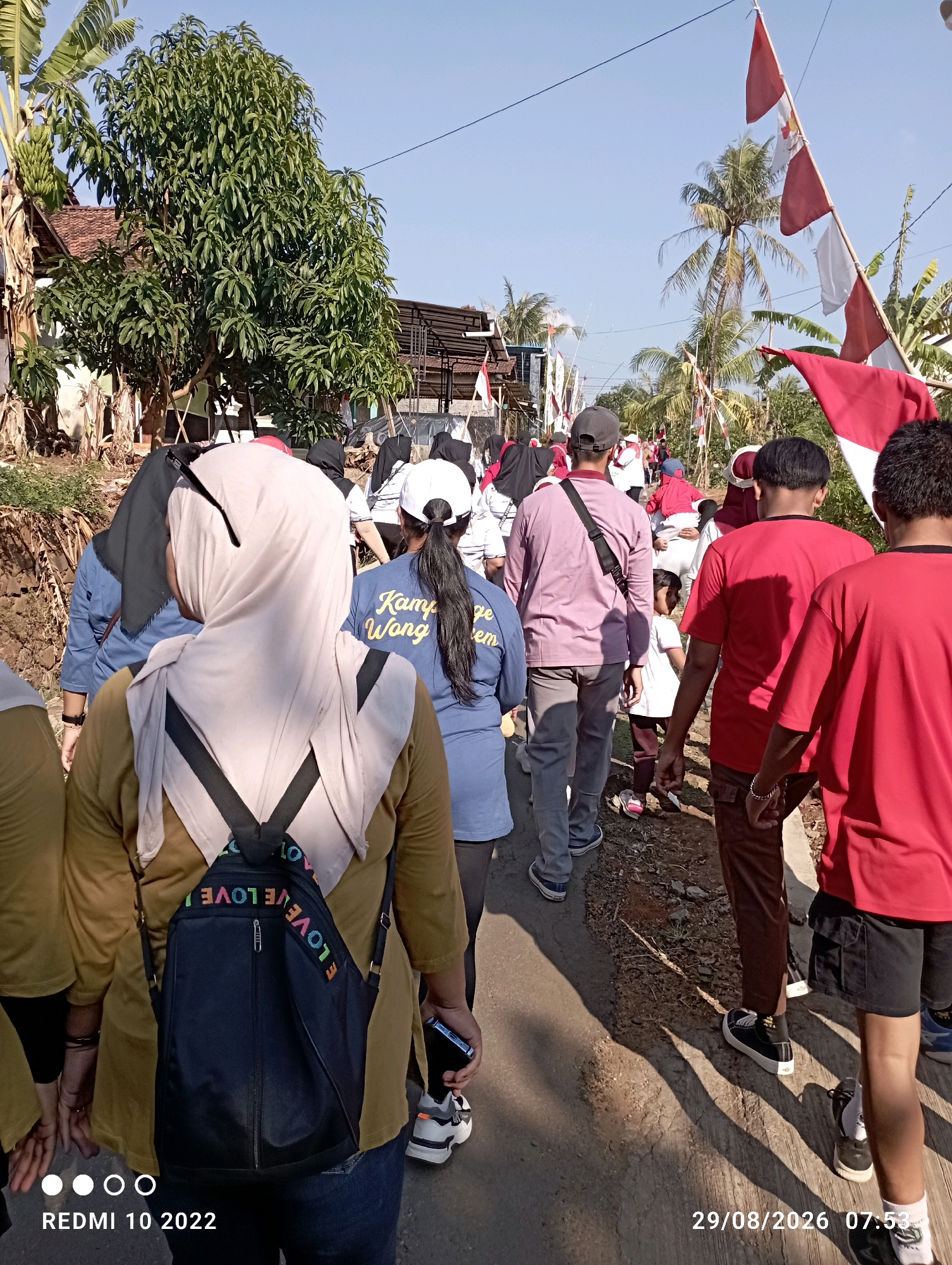 In Dukuhseti, Indonesia, on August 29, 2026, at 7:53 AM, a group of people are walking along a paved road lined with trees and buildings. The people are dressed in casual clothing, with some wearing red shirts and others wearing more subdued colors. Several individuals are wearing headscarves. Red and white flags, likely Indonesian flags, are visible in the background, suggesting a celebratory or national event is taking place. Some of the people are carrying backpacks, and one person is holding a mobile phone. The weather appears to be clear and sunny, with shadows cast on the road by the people and surrounding vegetation. The scene depicts a community gathering or procession in a rural or semi-rural setting.