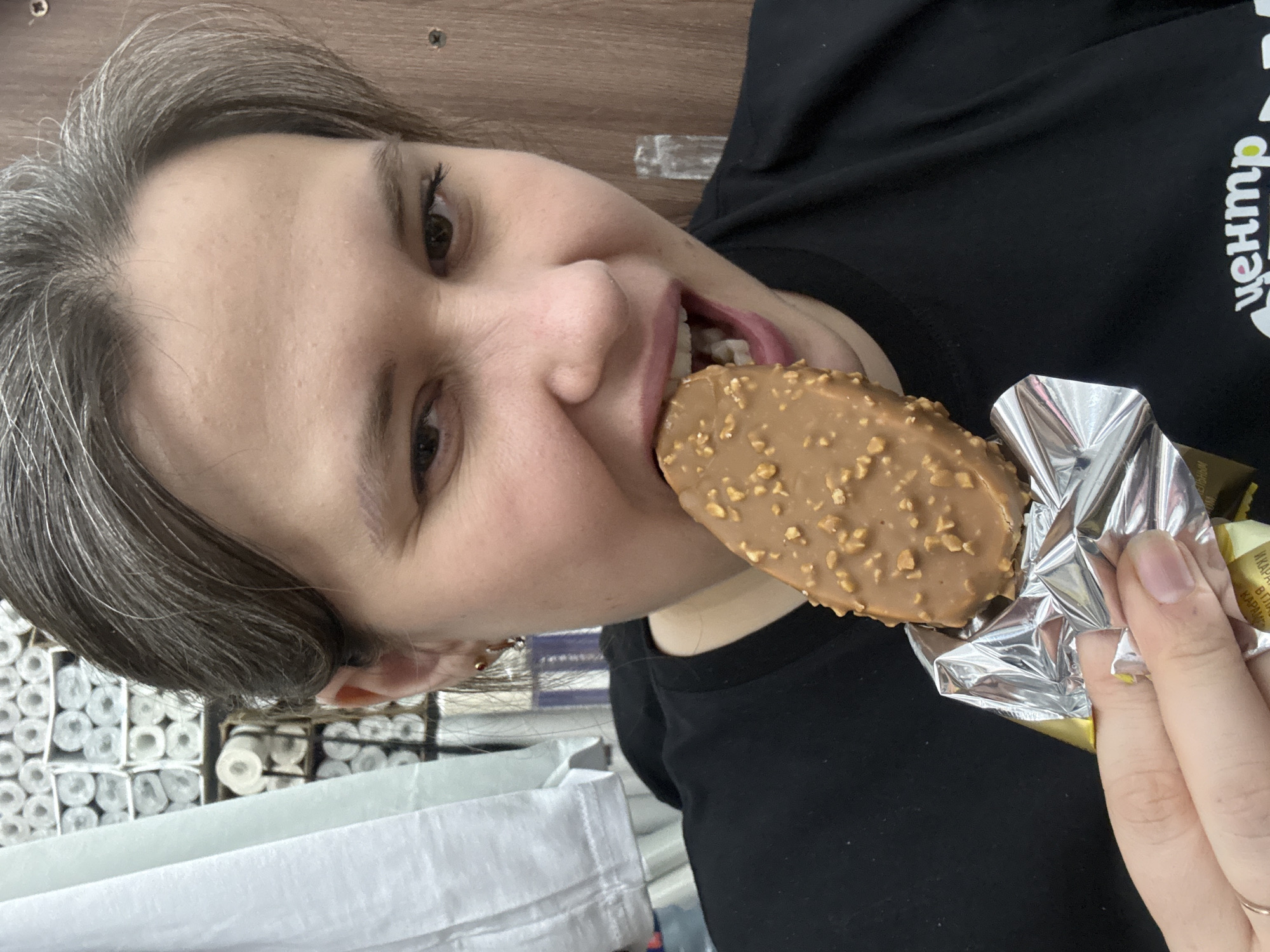 A woman with short grey hair is eating an ice cream bar covered in nuts. She is wearing a black t-shirt with white writing on it. The ice cream is partially unwrapped. Rolls of paper are in the background. The location is Chaikovsky, Russia.