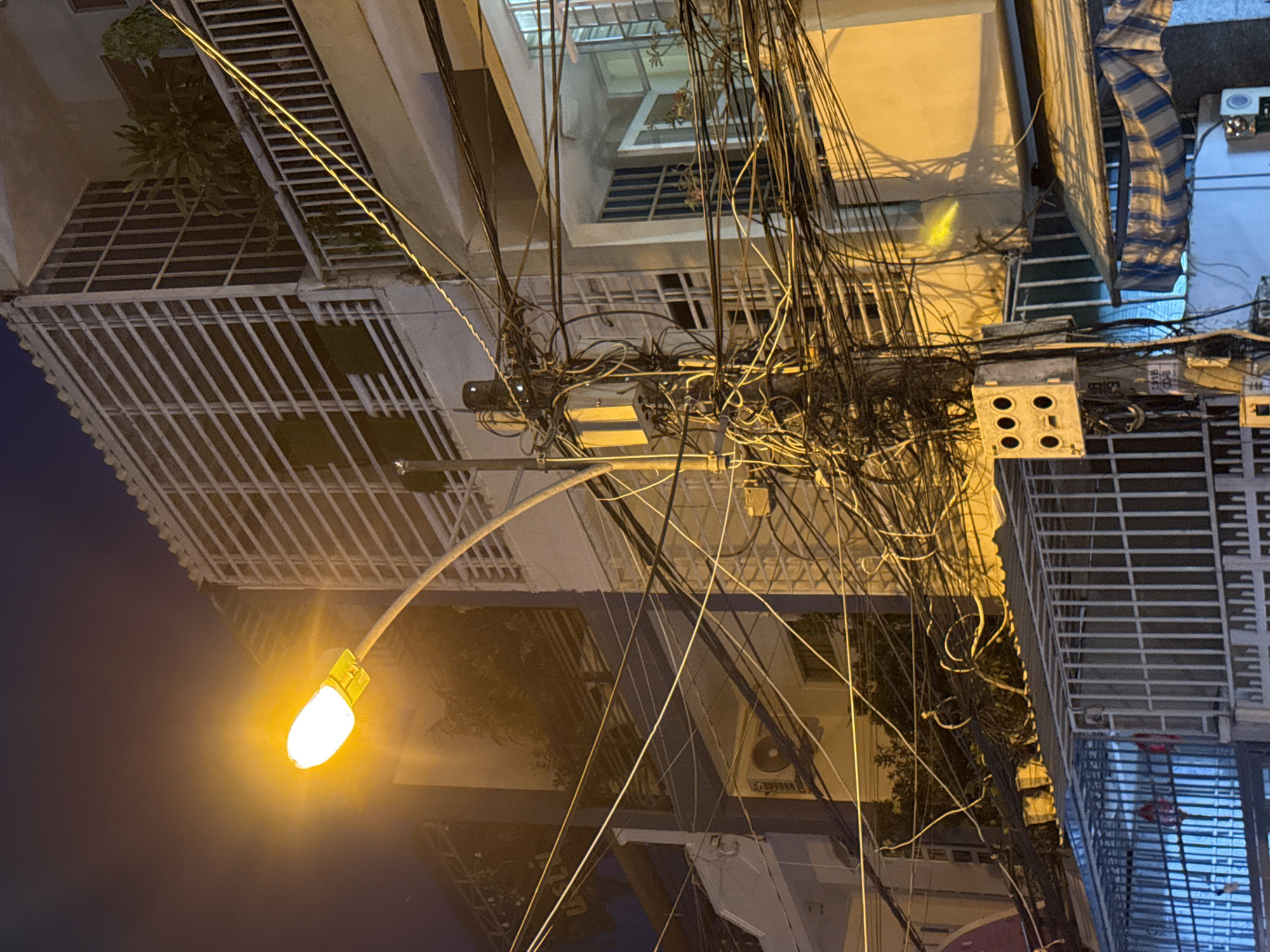 The image depicts a street scene at night in Nha Trang, Vietnam, characterized by a complex and dense network of overhead electrical and telecommunication wires. A brightly lit streetlamp illuminates the scene with a warm, orange glow. In the background, multi-story buildings with balconies and window grates are visible, some with lush green plants. A utility pole, heavily laden with wires, stands prominently in the foreground, equipped with electrical boxes and meters. A white metal fence runs across the bottom of the frame. The overall impression is one of urban infrastructure, typical of a developing city.