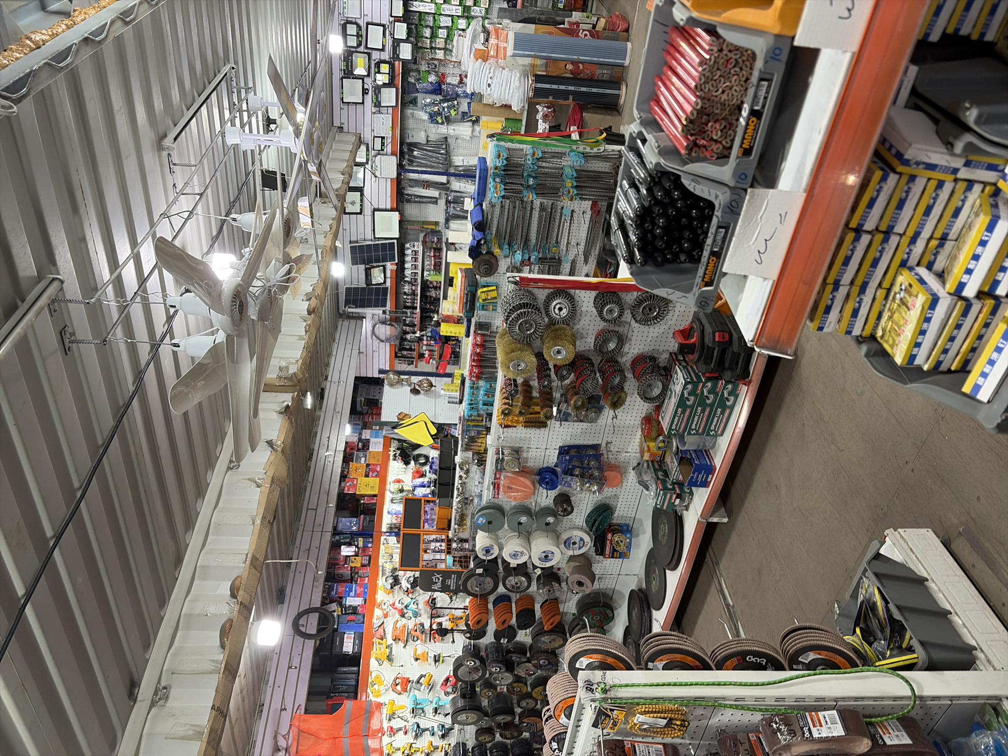 The image depicts the interior of a hardware store located in Kant, Kyrgyzstan. The store is filled with an extensive display of tools and hardware supplies. Various sections showcase items such as power tools, cutting discs, wire brushes, sanding equipment, lighting fixtures, and other related products. The shelves are densely packed, indicating a wide inventory. The store appears to be well-lit with overhead lighting. No individuals are visible in the image. The environment is organized with merchandise displayed on pegboards and shelves.