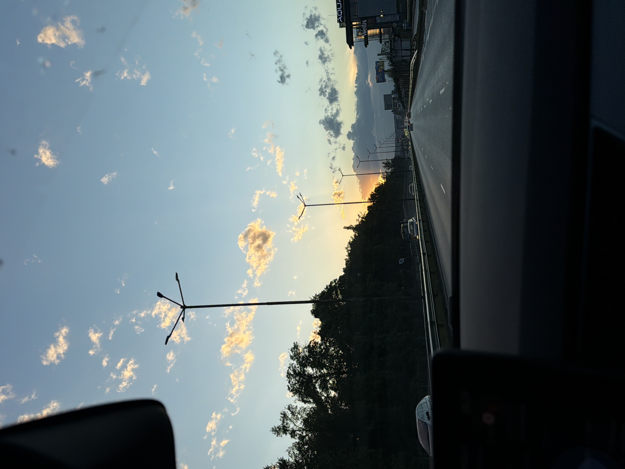 This media file captures an outdoor scene from inside a moving vehicle, rotated 90 degrees clockwise from its natural orientation. When viewed correctly, the image shows a blue sky with scattered, fluffy cumulus clouds, illuminated by the warm, golden light of what appears to be a sunset or sunrise on the left horizon.
The foreground on the right and bottom is dominated by the dark interior elements of the vehicle, providing a natural frame. The main subject is a multi-lane road or highway stretching into the distance on the right, lined with various elements. White lane markings are visible on the asphalt. Several vehicles can be seen on the road, receding into the distance, some identifiable by their faint headlights or taillights.
Two prominent, modern streetlights with multiple arms are silhouetted against the bright sky and the setting sun. A dark, dense treeline or forest runs along the left side of the road, also silhouetted against the sky's glow. In the far distance on the right, beyond the road and trees, urban buildings are visible. One building clearly displays the text "AQUA" and "WELLS" on signs.
The setting is an urban or suburban area, likely a major road or highway in Sofia, Bulgaria, consistent with the visible commercial signs. The time of day is either late afternoon nearing sunset or early morning during sunrise, given the low sun and the golden hues painting the clouds and horizon. The weather appears clear with light cloud cover. No people are explicitly visible, but the scene implies activity of driving.