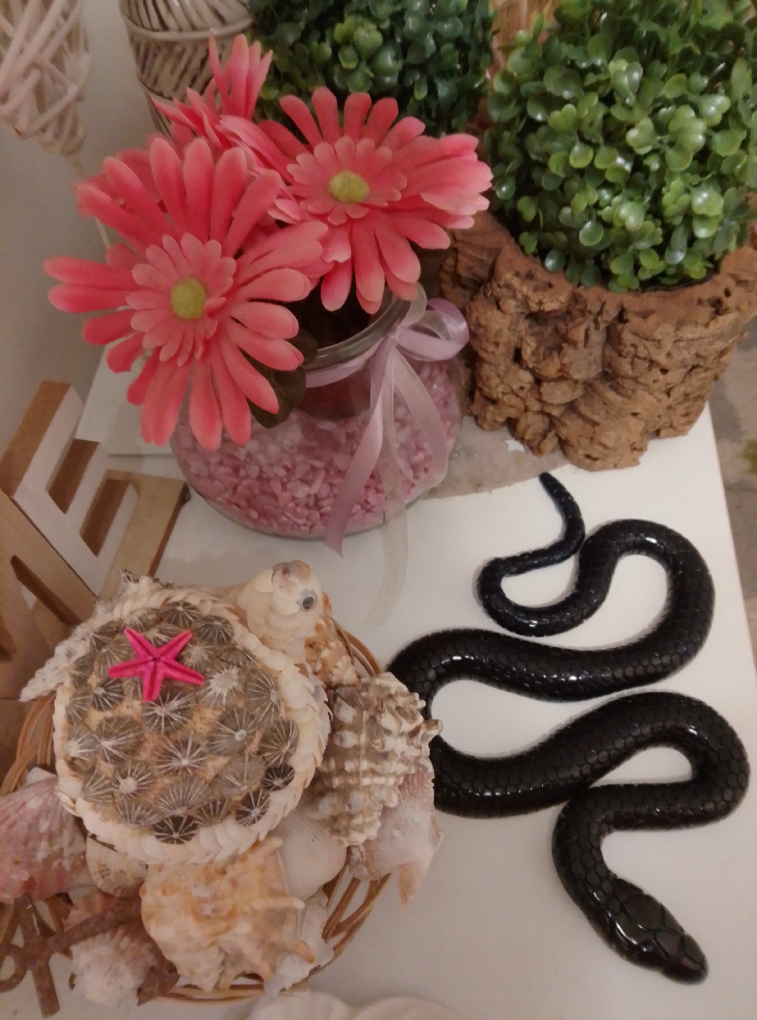 A decorative display featuring a black snake figurine is shown on a white surface. To the left of the snake, a basket overflows with various seashells, and prominently displayed on top of the shells is a circular object adorned with spiky patterns and topped with a pink starfish. Behind the basket, a glass jar filled with pink pebbles is visible, with a cluster of pink artificial flowers emerging from it. Further to the right, a rough-textured brown pot contains a lush green artificial plant. The scene appears to be a still life arrangement, possibly for decorative purposes. No people are present in the image, and there is no specific information to indicate its location in Wedel, Germany.