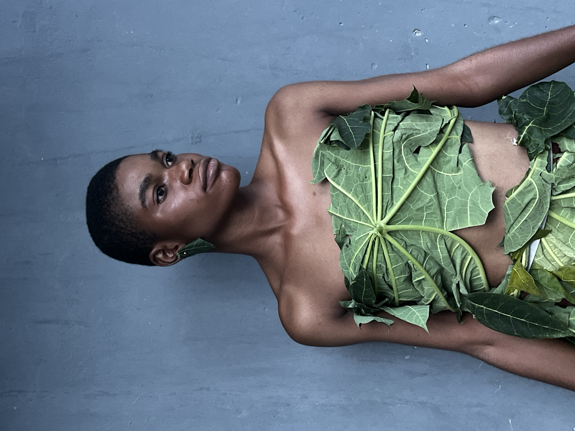 A brown-skinned person is lying on their back on a gray concrete surface. They are partially covered by large green leaves. They are oriented with their head tilted upwards and towards the left, and their body extending downwards to the right. A single green leaf is placed as an earring on their left ear. The person's head is closely shaved. The background is a plain gray color, possibly a painted wall or floor. The location, while potentially in the city of Badagry, Nigeria, cannot be confirmed from the image alone.