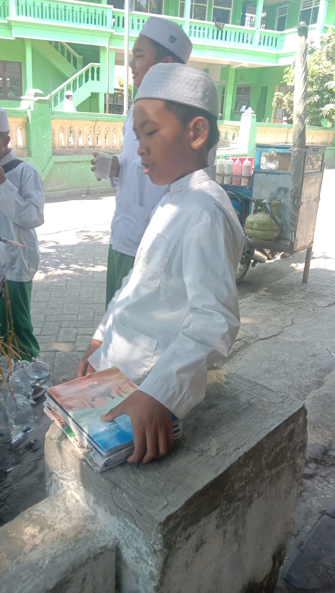 In Ngempit, Indonesia, on a sunny day, a young boy wearing a white tunic and a traditional cap stands at the foreground of the image. He is holding a stack of books on a concrete structure. His right hand is on top of the books. Behind him, another boy in similar attire is visible, holding a cup. In the background, a green building with multiple floors is present, along with trees and a street vendor's cart equipped with bottles and a gas cylinder. The ground appears to be paved with bricks. The scene suggests a school or religious institution setting, possibly during a break or after an event.
