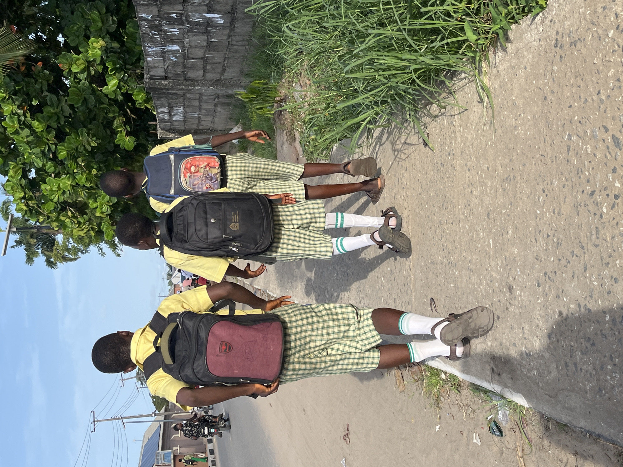 Three school children in yellow shirts, green and white plaid shorts, and white socks with green stripes are walking away from the viewer on a sidewalk. Each child wears sandals and carries a backpack. The first backpack is maroon and black, the second is black, and the third is blue with cartoon characters. To the right, a patch of grass grows near the sidewalk. The street on the left is paved and shows a motorcycle in the distance, utility poles with wires, and buildings. The background includes a cloudy blue sky. The location appears to be in an urban area, potentially Badagry, Nigeria.