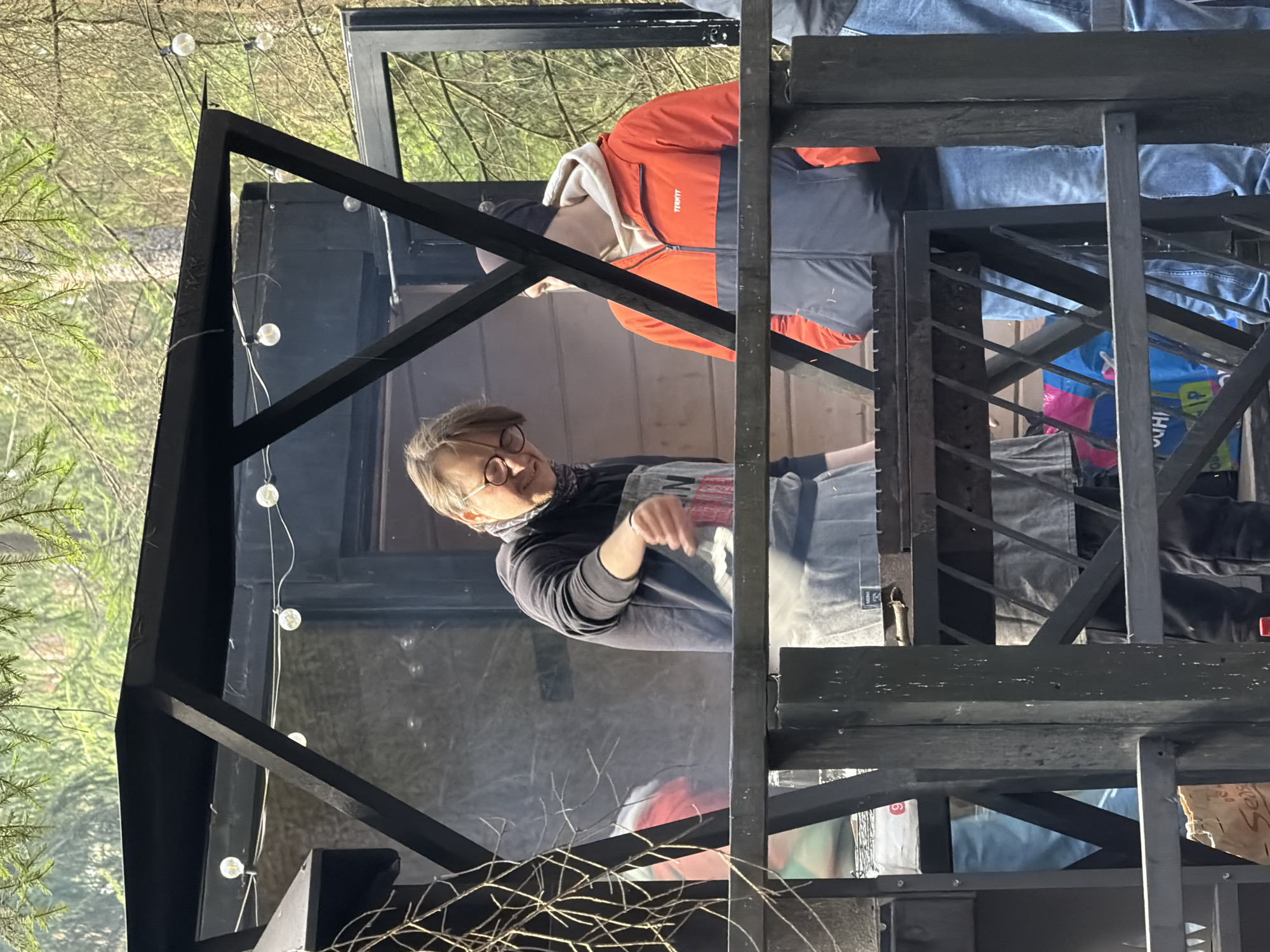 The image depicts a multi-level wooden structure with a black frame, possibly a stage or installation, located outdoors in Vsevolozhsk, Russia. The background shows a wooded area with green trees. String lights are attached to the structure. A man with glasses stands on one level, and another person wearing an orange and gray jacket is visible on a higher level. Miscellaneous objects, including bags and other items, are stored or displayed on the structure.