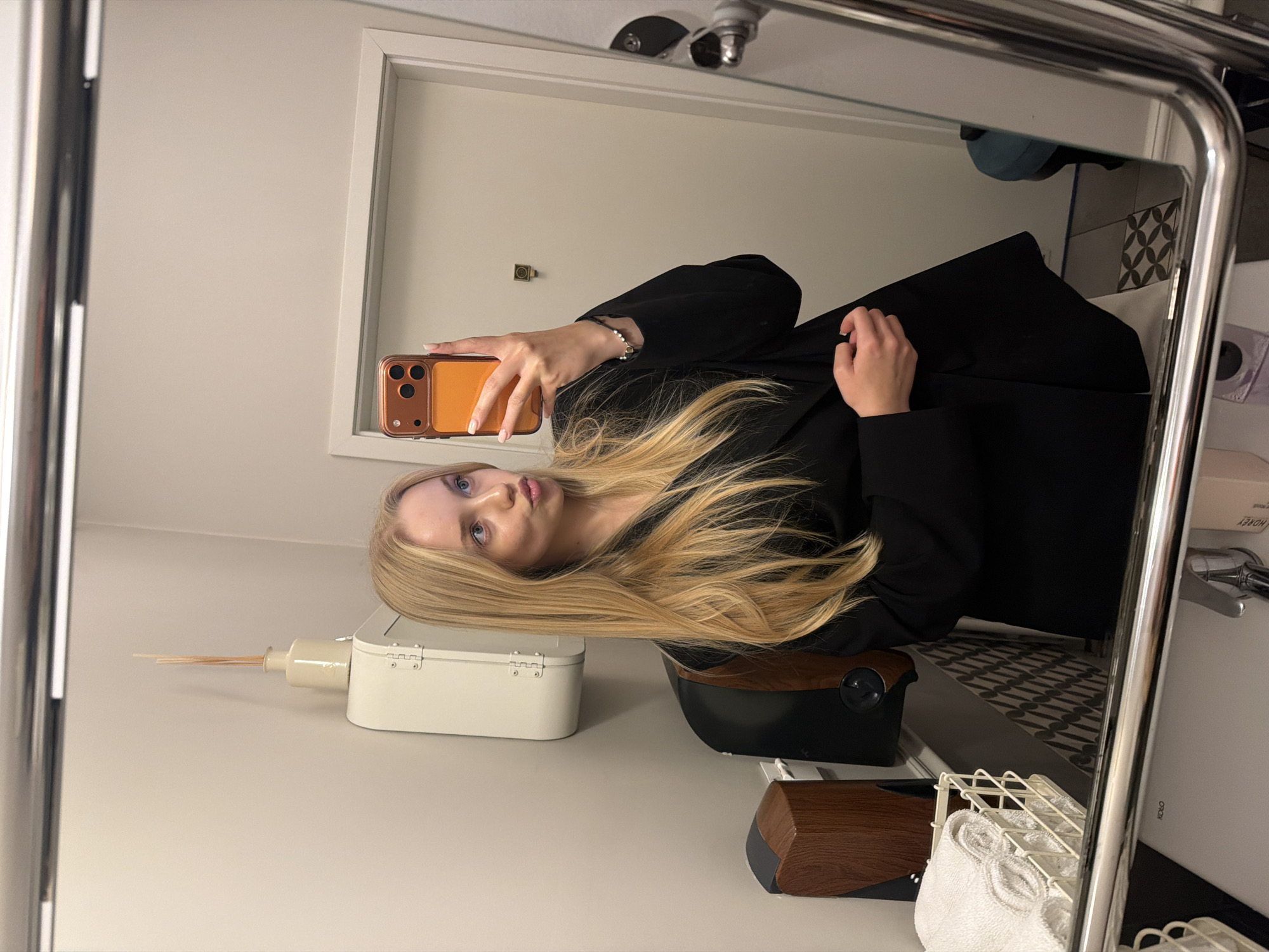 A mirrored selfie of a blonde-haired woman in a black coat is captured in a bathroom setting, likely in Unknown, Unknown. The woman holds a phone with an orange and brown case at eye level, partially obscuring her face. She has blue eyeshadow and fair skin. Her long, blonde hair is styled in loose waves. The bathroom features a white cabinet with a dispenser of wooden sticks next to it, a black chair, and a dark wood and black soap dispenser. In the background, a white sink countertop with a wire basket holding white towels is visible, alongside decorative patterned tiles and a blue object possibly indicating a shower or bath area. A framed mirror is also visible in the mirror's reflection.