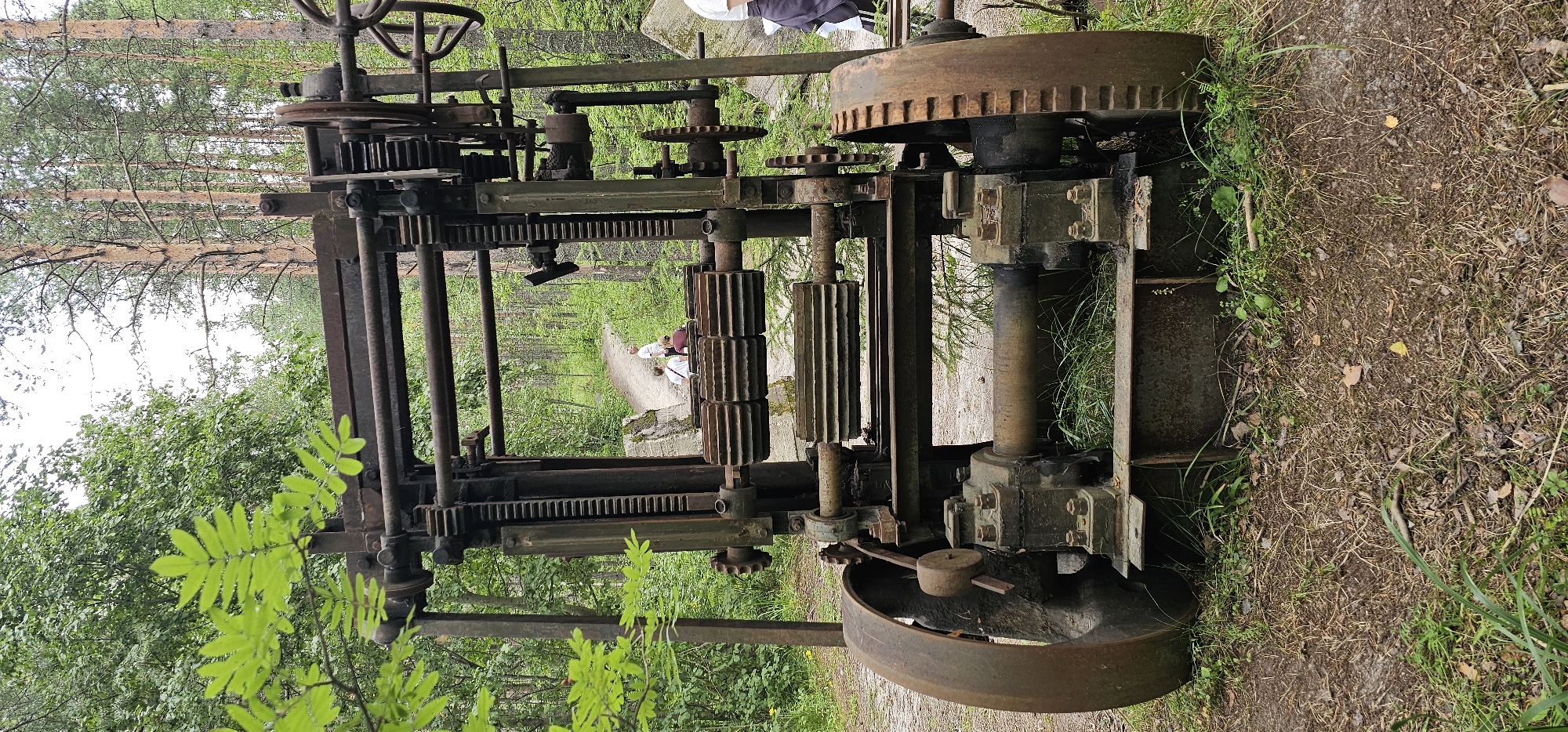 In Medvezhyegorsk, Russia, an abandoned industrial machine, likely from the Soviet era, rests amidst a forest. The machine, characterized by its large gears, rusted metal frame, and weathered components, stands on a dirt path. The surrounding environment is a dense forest with tall pine trees and lush green foliage. In the background, two individuals are visible walking along the path, adding a sense of human presence to the scene.