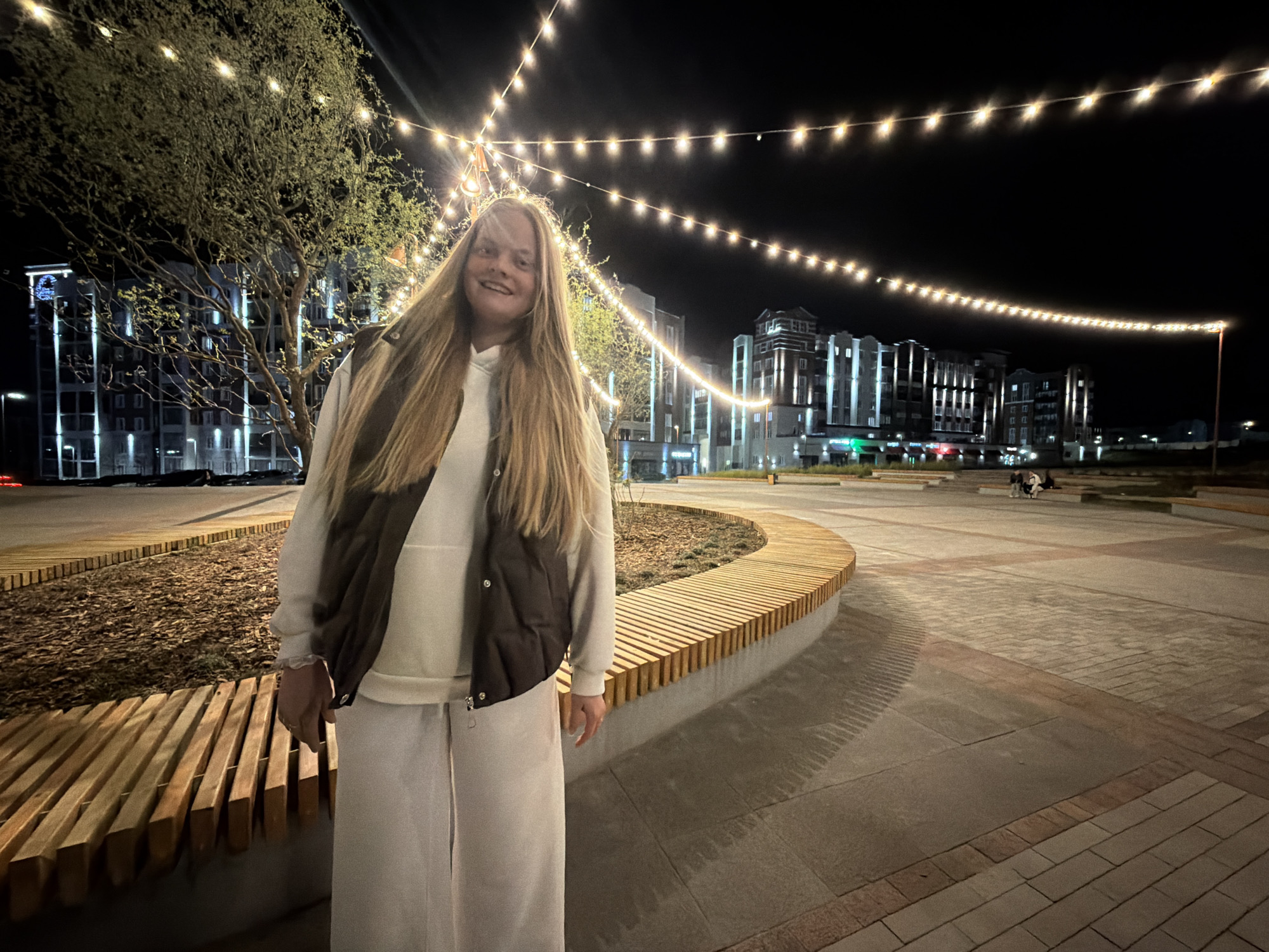 A young, fair-skinned woman with long, blonde hair stands on a paved walkway in Izhevsk, Russia, at night. She is wearing a white sweatshirt, white sweatpants, and a dark brown vest. A tree is to her left, and a curved wooden bench is behind her. In the background, buildings are visible, illuminated with white lights, along with strings of decorative lights strung overhead. Other people are visible in the background, sitting on benches. The walkway is paved with rectangular tiles. The lighting suggests it is nighttime.