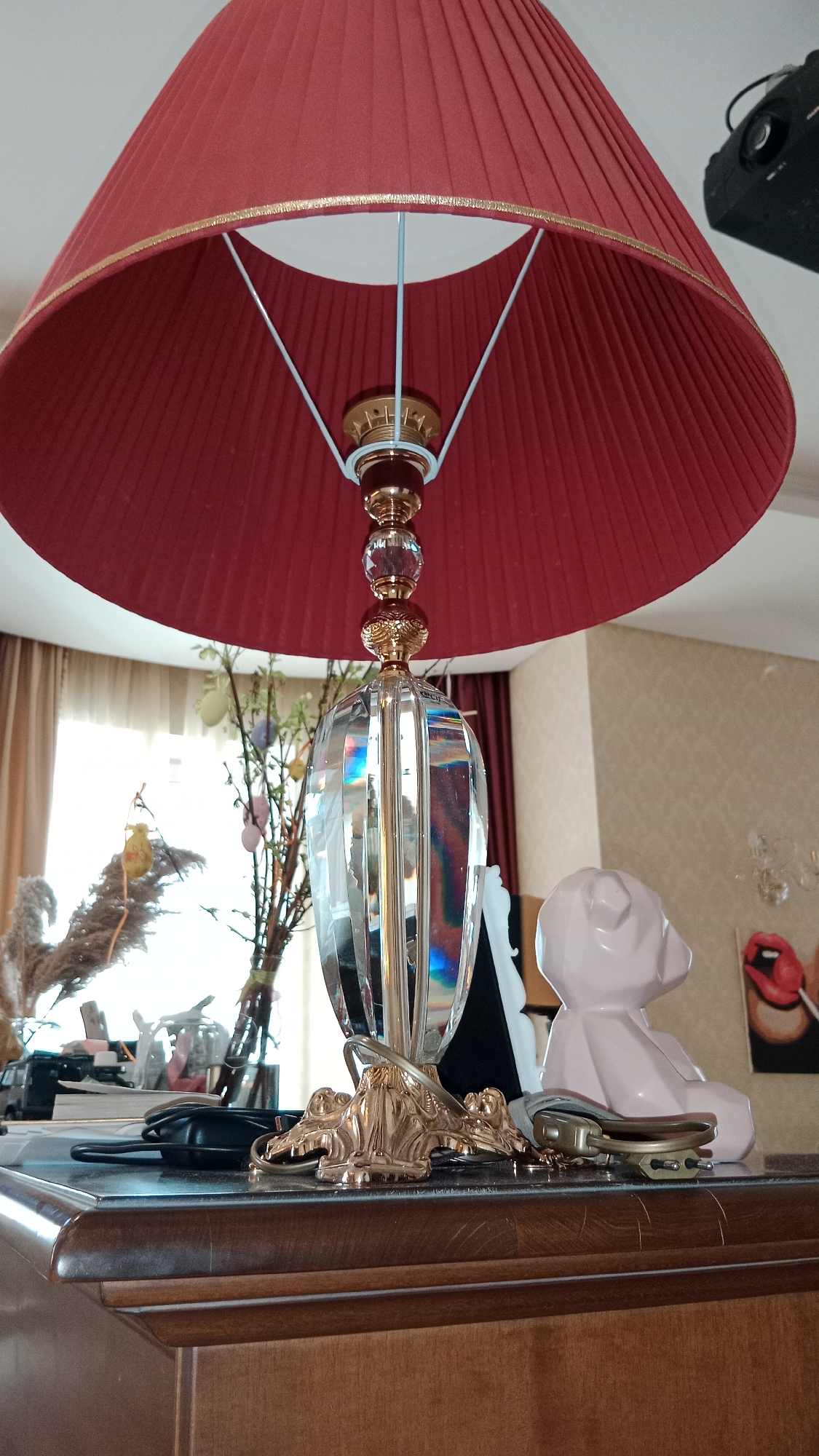 A low-angle, close-up shot of a table in what appears to be a residential interior. Dominating the foreground is a large lamp with a pleated red lampshade, trimmed with a gold-colored accent. The lamp's body is a multifaceted glass form mounted on an ornate gold base, with a finial consisting of gold and crystal-like spheres.
The table's surface is cluttered. A white ceramic bear-shaped figurine sits to the right of the lamp. Behind the lamp, near a window, there are branches decorated with Easter eggs in a glass vase. Further back, artwork with red lips is visible. A black object is positioned between the lamp and the white figurine, partially obscured. Other miscellaneous items, including electronic devices, are also present on the table.
The lighting is bright, natural light emanating from a window to the left of the frame. The location is likely a private residence. There is no explicit evidence to suggest the location is Moscow, Russia.