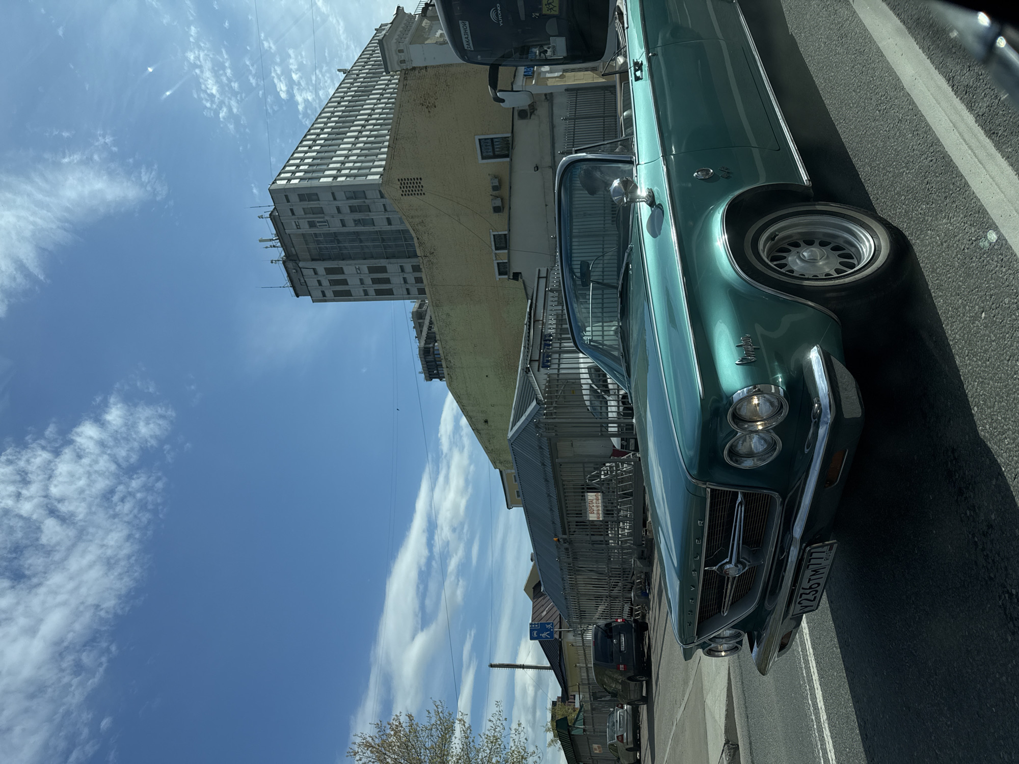 The image shows a teal vintage Chrysler car parked on the side of a street. The car is oriented at an angle, with its front facing the bottom of the image. Visible details include the front grill, headlights, and license plate.
In the background, a tall, multi-story building with a grid-like facade is visible, partially obscured by other buildings. A blue sky with white clouds forms the backdrop.
Based on the license plate visible on the car, the location appears to be Moscow, Russia.