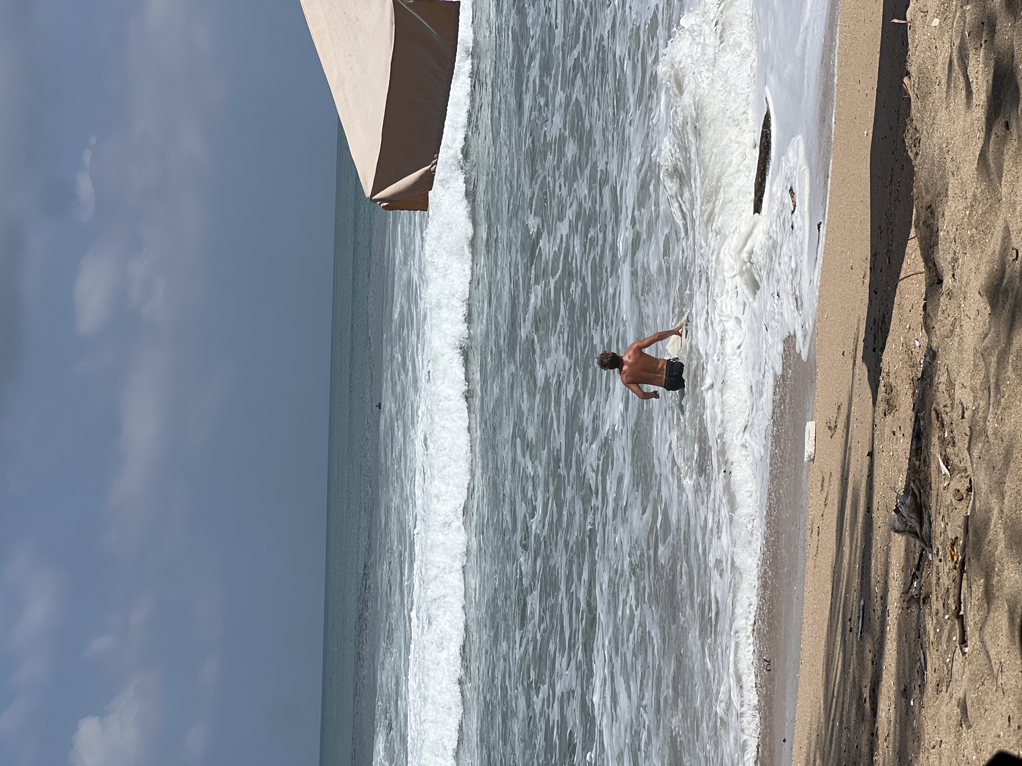A shirtless person, facing away from the camera, walks into the ocean at Kuta Beach, Indonesia. The individual is wearing dark-colored shorts and holding a surfboard. The water is shallow around their legs, with waves breaking and creating white foam as they approach the shore. The beach is sandy, with some debris scattered near the waterline. In the background, the ocean stretches to the horizon under a cloudy sky, and a portion of a large, light-colored umbrella is visible in the upper right corner of the frame.