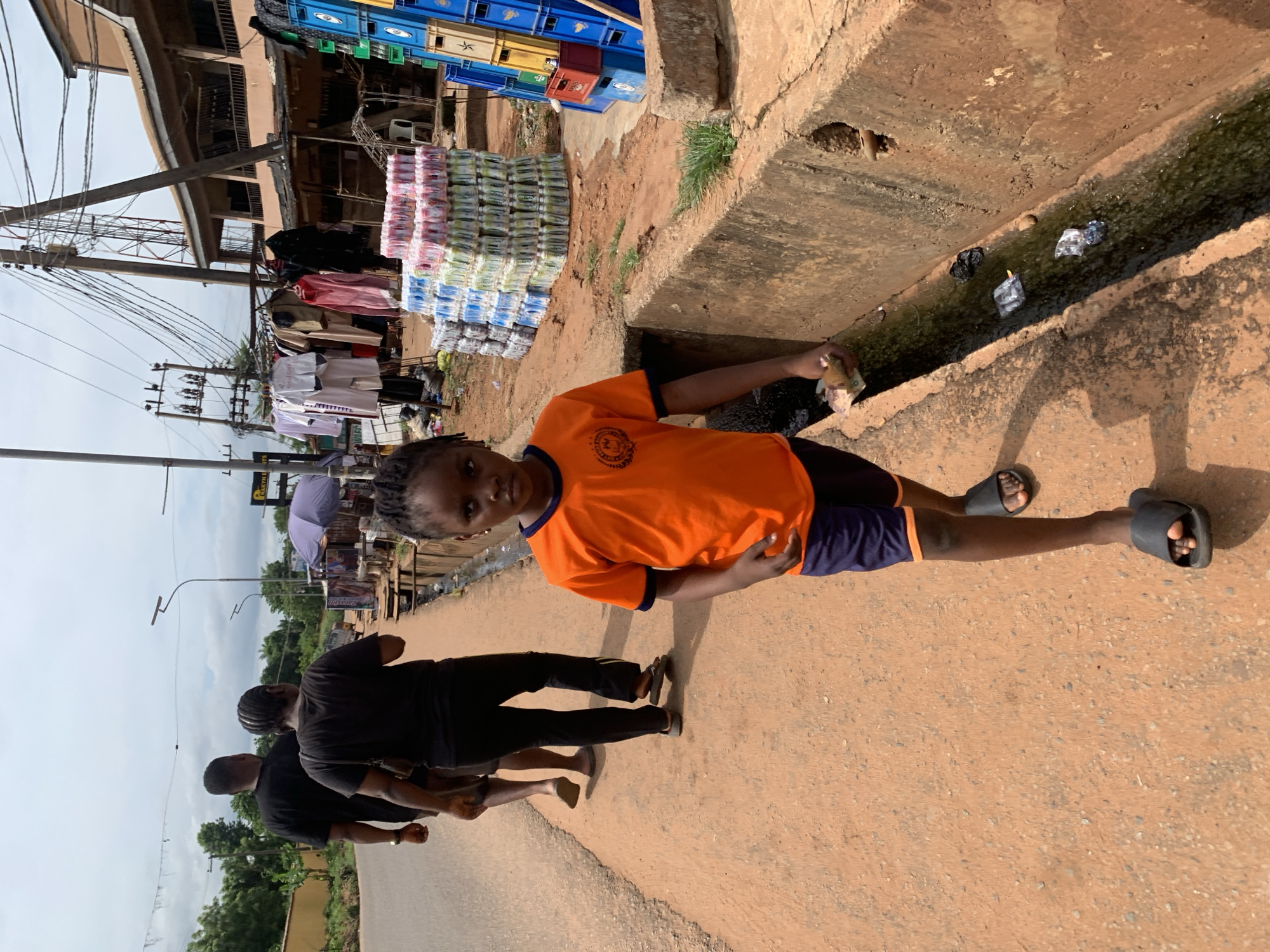 The image depicts a girl in an orange t-shirt standing next to a concrete drainage ditch on a paved surface in what appears to be the city of Ukwu-Abwa, Nigeria. She is wearing dark shorts and sandals, and she holds a crumpled object in her hand. The drainage ditch contains water and some debris. Behind her, two people in dark clothing walk along the pavement. Stacks of packaged goods and colorful crates are visible in the background, along with buildings and power lines.