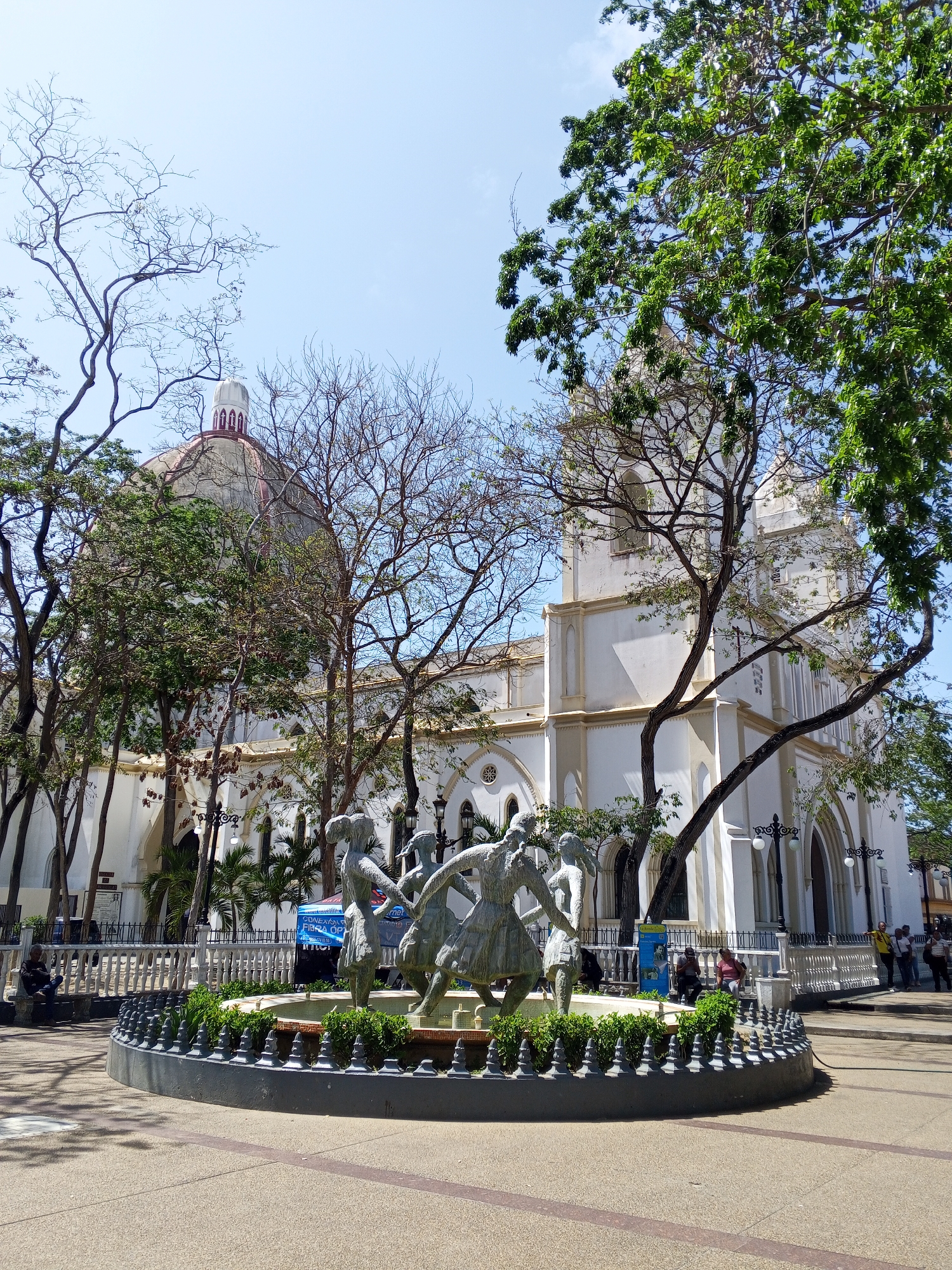 The image captures a bright, sunny day in Porlamar, Venezuela, featuring the main square, Plaza Bolívar. A prominent white church stands in the background, partially obscured by trees, with a dome visible on the left. In the foreground, a circular fountain is adorned with a bronze statue of children dancing. People are scattered throughout the square, some sitting on benches, others walking. The ground is paved, with trees providing shade. The overall atmosphere is relaxed and public.
