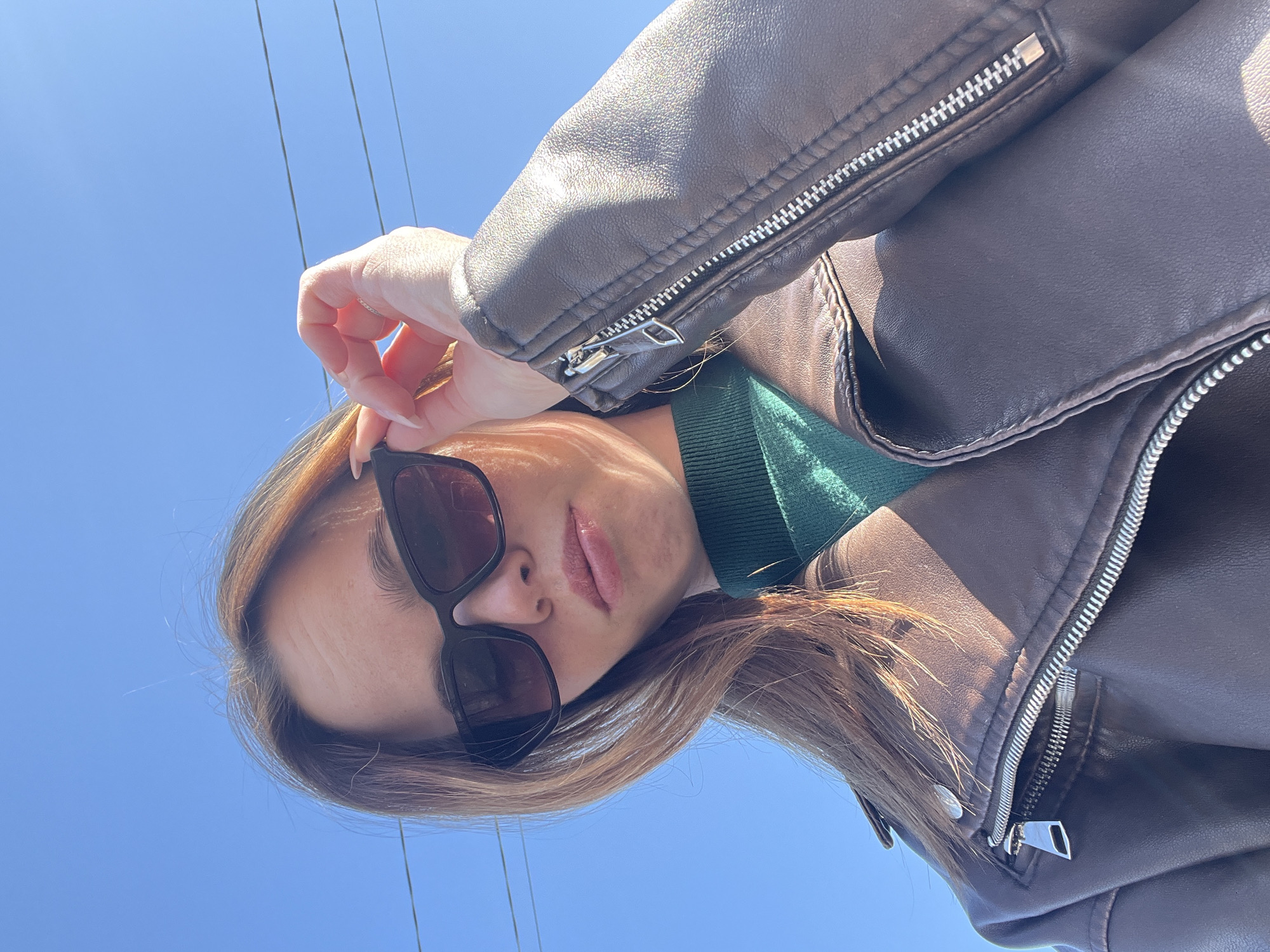 A woman wearing sunglasses, a green turtleneck sweater, and a brown leather jacket stands outdoors. The backdrop is a clear, bright blue sky. Power lines are visible in the upper left corner. The photograph seems to have been taken from a low angle, looking up at the subject. The location is Chaikovsky, Russia.