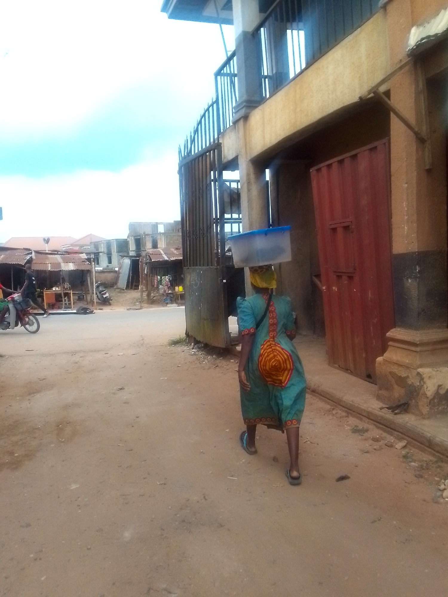 A woman wearing a blue dress with a circular design on the back walks away from the camera. She has a light blue plastic container on her head, and is wearing sandals. She is walking on a dirt road next to a building with a red metal door and a wrought iron gate. In the background, there is a street with shops and a man on a motorcycle. The scene is in Orlu, Nigeria.