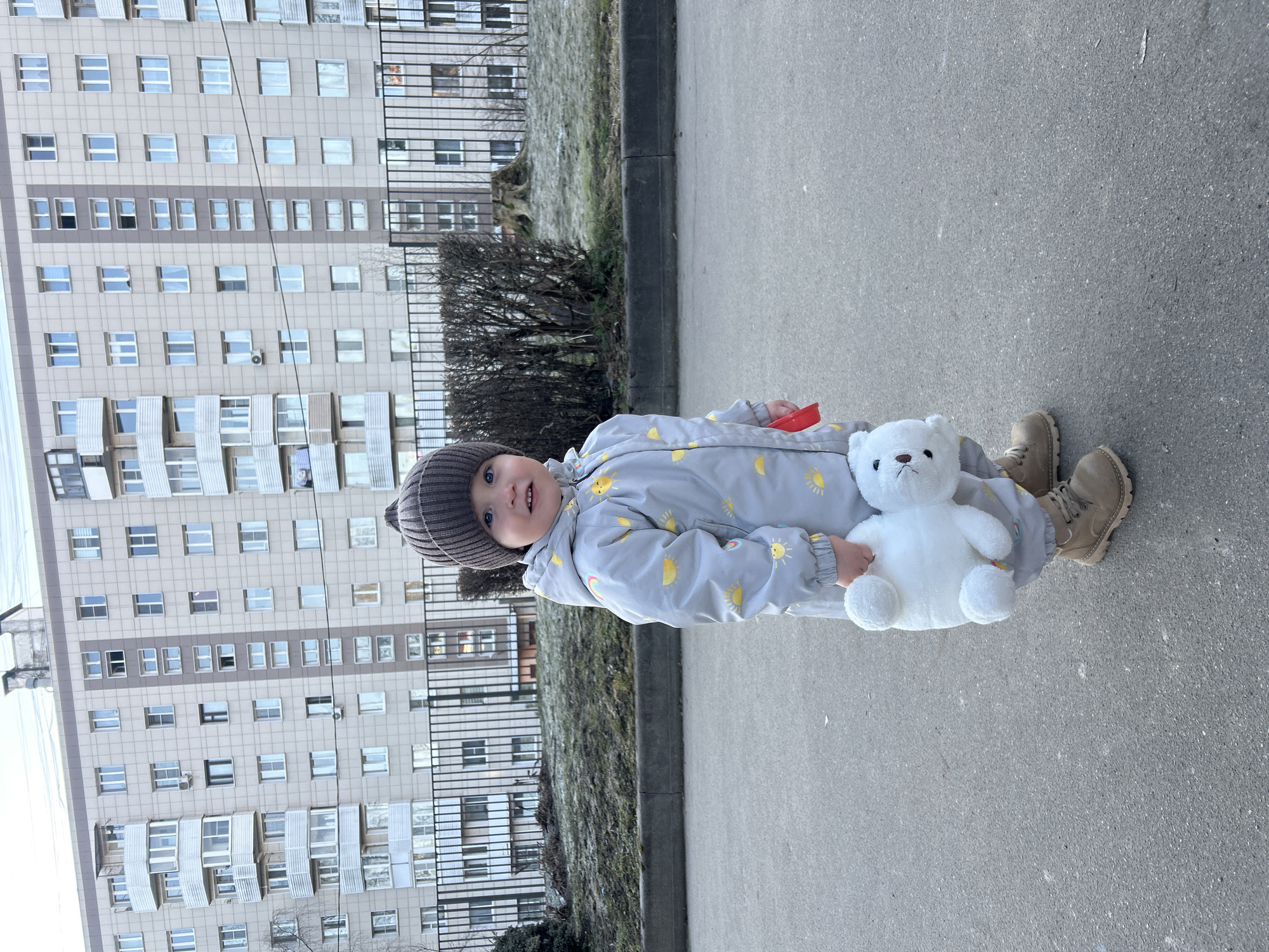 A young child stands on an asphalt surface next to a curb and a grassy area with a bush. The child wears a grey snowsuit with yellow sun patterns and a brown knit hat, and tan boots. The child is holding a white teddy bear and a red toy. Behind the child are a black metal fence and a large, multi-story residential building. The location is in the city of Klin, Russia.