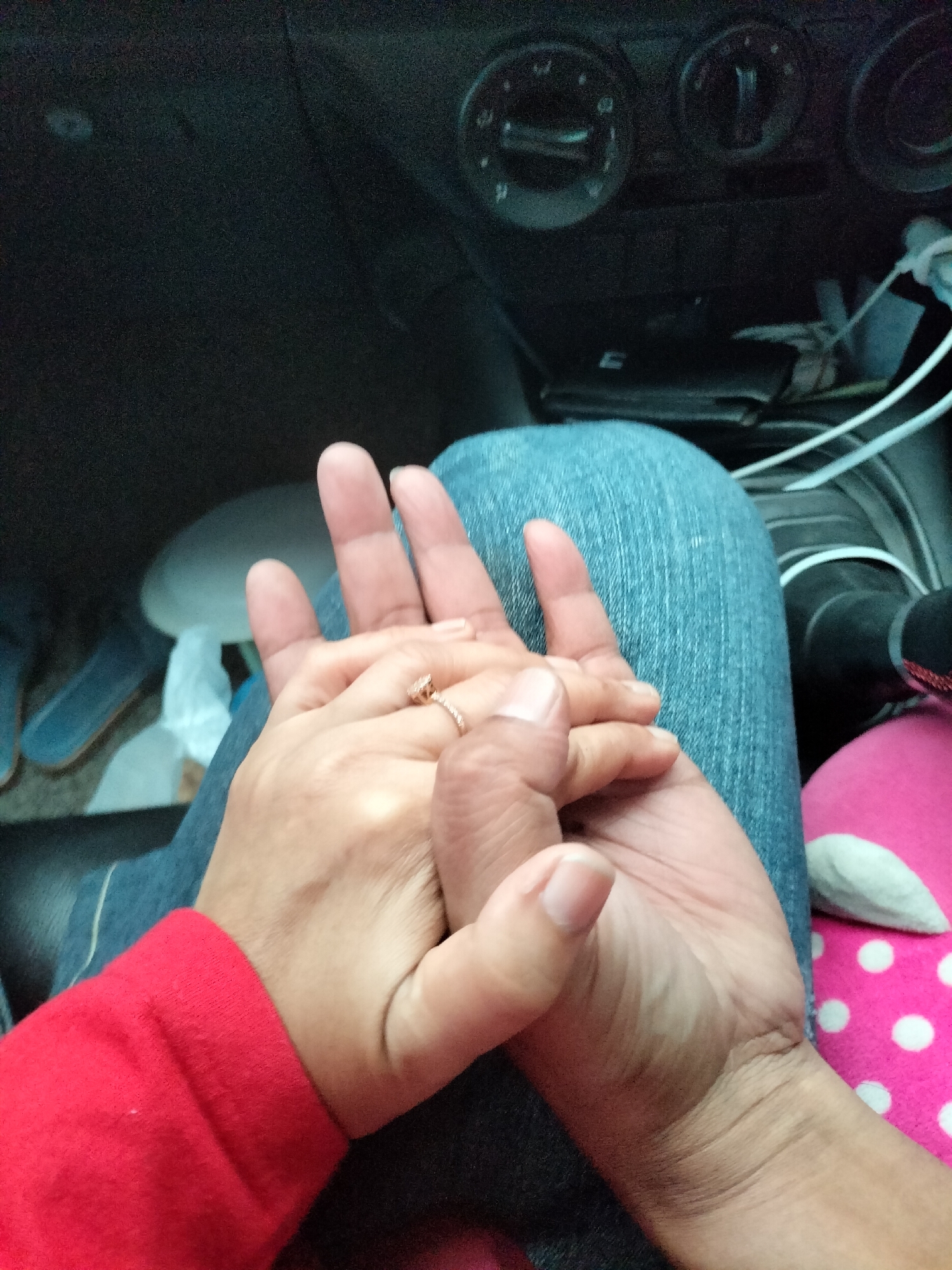 Two individuals are holding hands in the passenger seat of a car. The person on the left is wearing a red long-sleeved shirt. The person on the right is wearing blue jeans. The hands are intertwined, and a ring is visible on one of the fingers. The background shows parts of the car's dashboard and interior. The scene is dimly lit. The location is identified as Bukit Selamat, Indonesia.