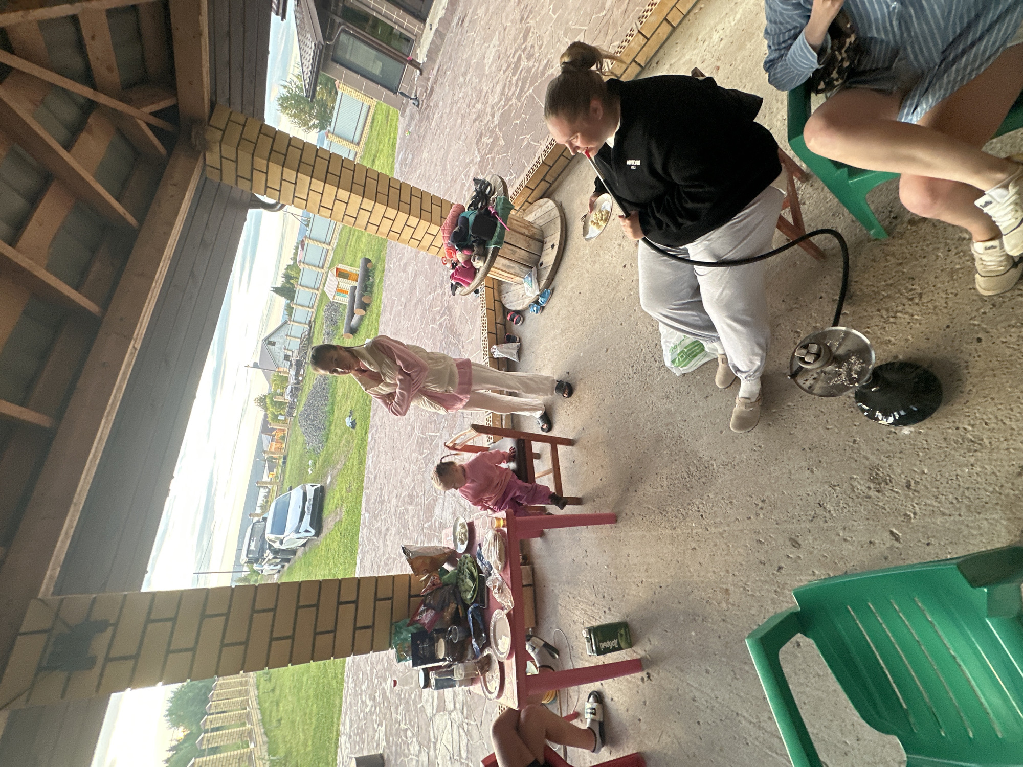 In the city of Peplayaeva, Russia, under a wooden-roofed open-air structure with brick pillars, a group of people are gathered. In the foreground, two individuals are seated on chairs, one of whom is smoking a hookah. To the left, a small pink table is laden with food and drinks, attended by a young child seated on a wooden chair. Further back, a woman stands with her arms crossed, looking towards the left. The ground is paved with concrete. In the background, a green lawn stretches out towards a fence, with houses and trees beyond. Two cars are visible parked on a dirt road. The sky is overcast.