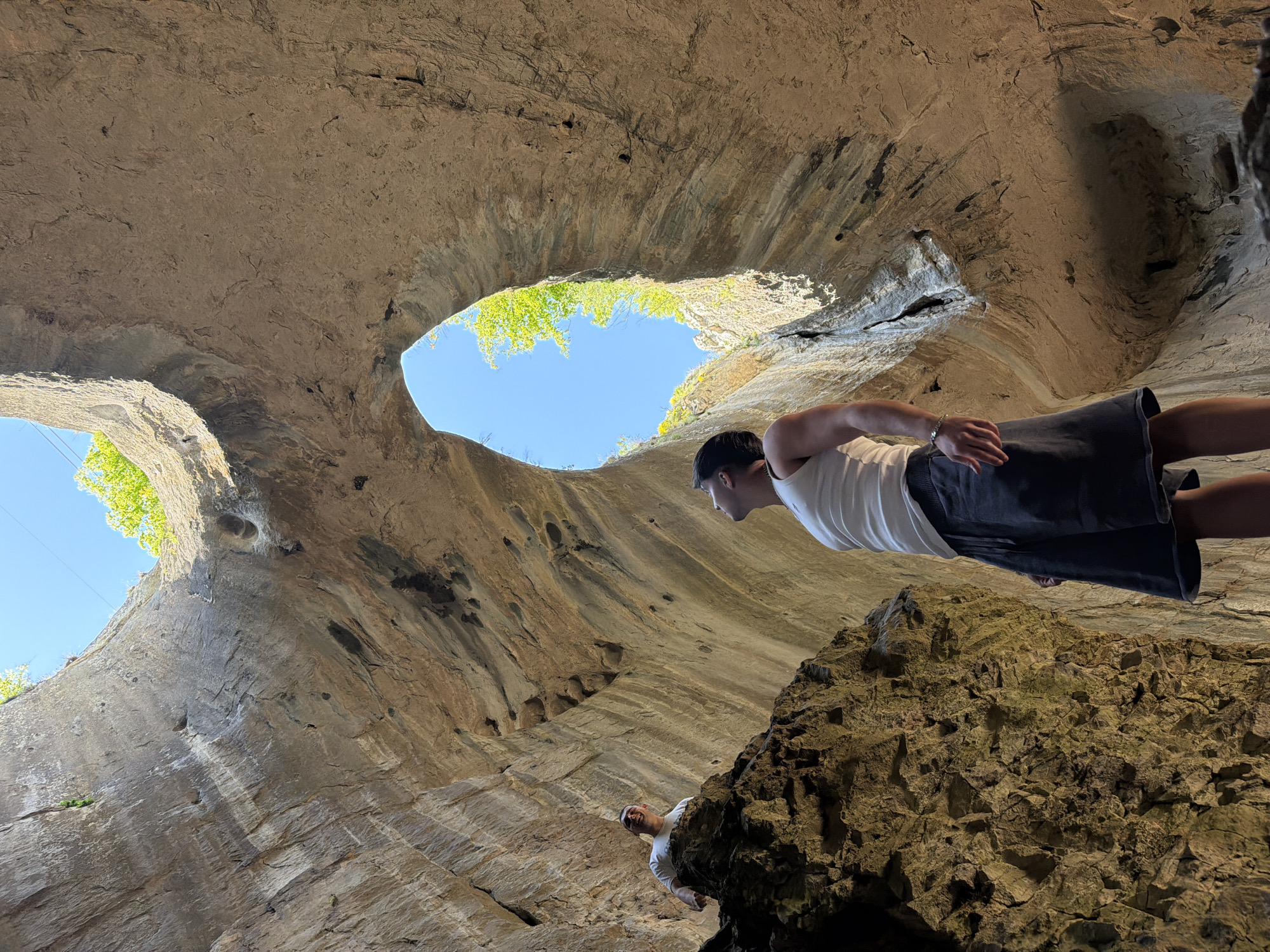 The image, taken in Karlukovo, Bulgaria, shows a man standing near the edge of a cave entrance. The cave has two large openings that reveal the sky and foliage. The man is wearing a white shirt and dark shorts. Another person is standing closer to the cave floor. The perspective is upward, emphasizing the cave's structure. A large rock formation is also visible.