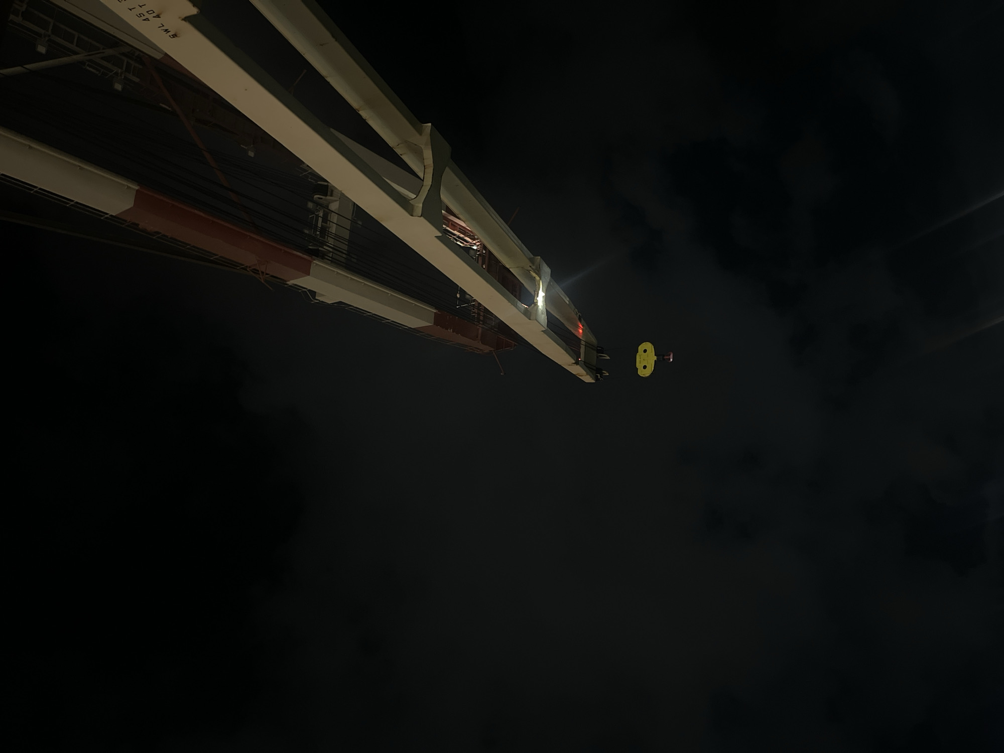 The image depicts a low-angle night shot of a large crane structure with a yellow object attached to the end of it, suspended against a dark sky with a few visible clouds. The crane is primarily white and red, and has a metal framework with numerous cables and structural supports. There is a light source illuminating the area around the end of the crane where the yellow object is attached. The location is in the city of Unknown, Unknown.