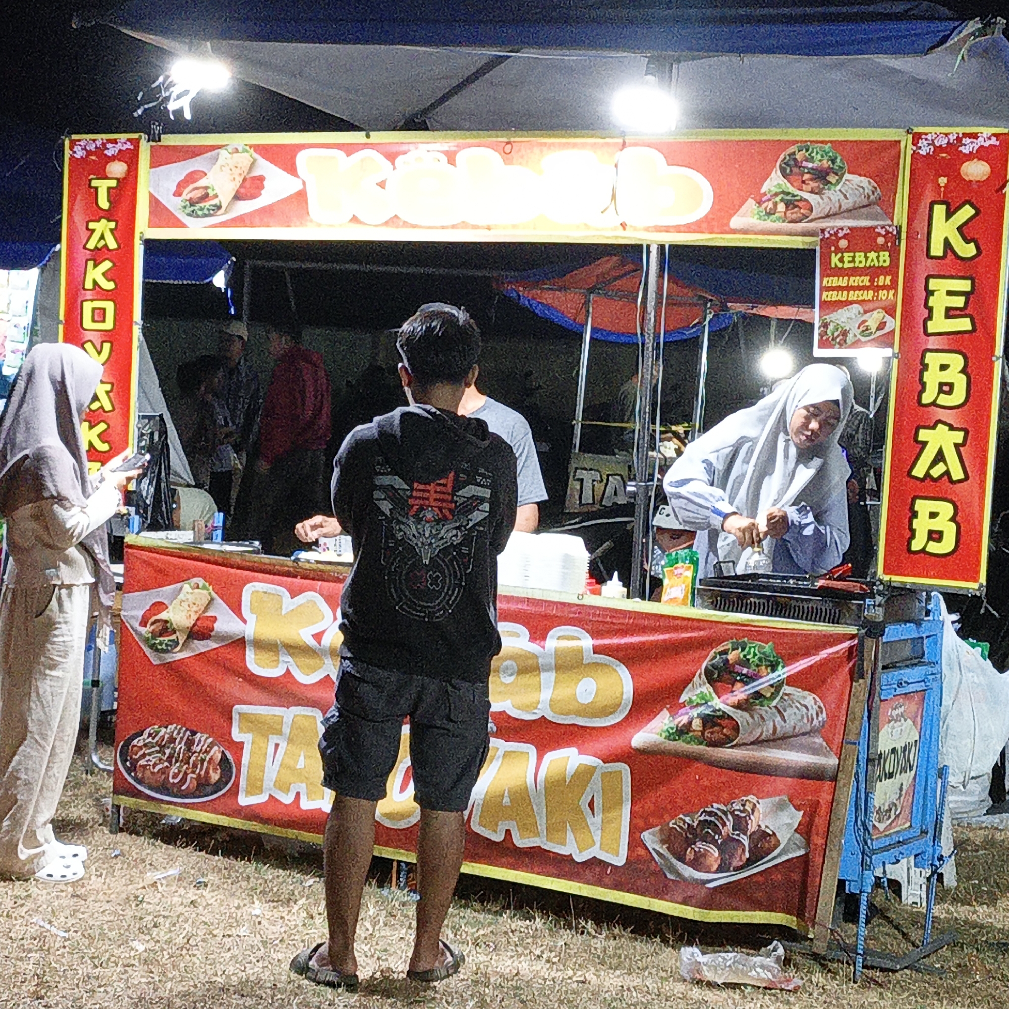 In Mangli, Indonesia, a food stall operates under a blue tent at night, illuminated by overhead lights. The stall displays a prominent red banner featuring images of kebabs and a plate of takoyaki. The stall's name, "TAKOKYA," is visible on the left. A woman in a light-colored hijab and pants stands on the left, holding a phone. On the right, a man wearing a black hoodie with a graphic design and dark shorts stands facing away from the camera. Another person in a white hijab works at the stall, preparing food. Behind them, other individuals are partially visible. The ground is covered in dry grass, with a discarded plastic bag in the foreground. The scene depicts a busy street food vendor in a community setting.