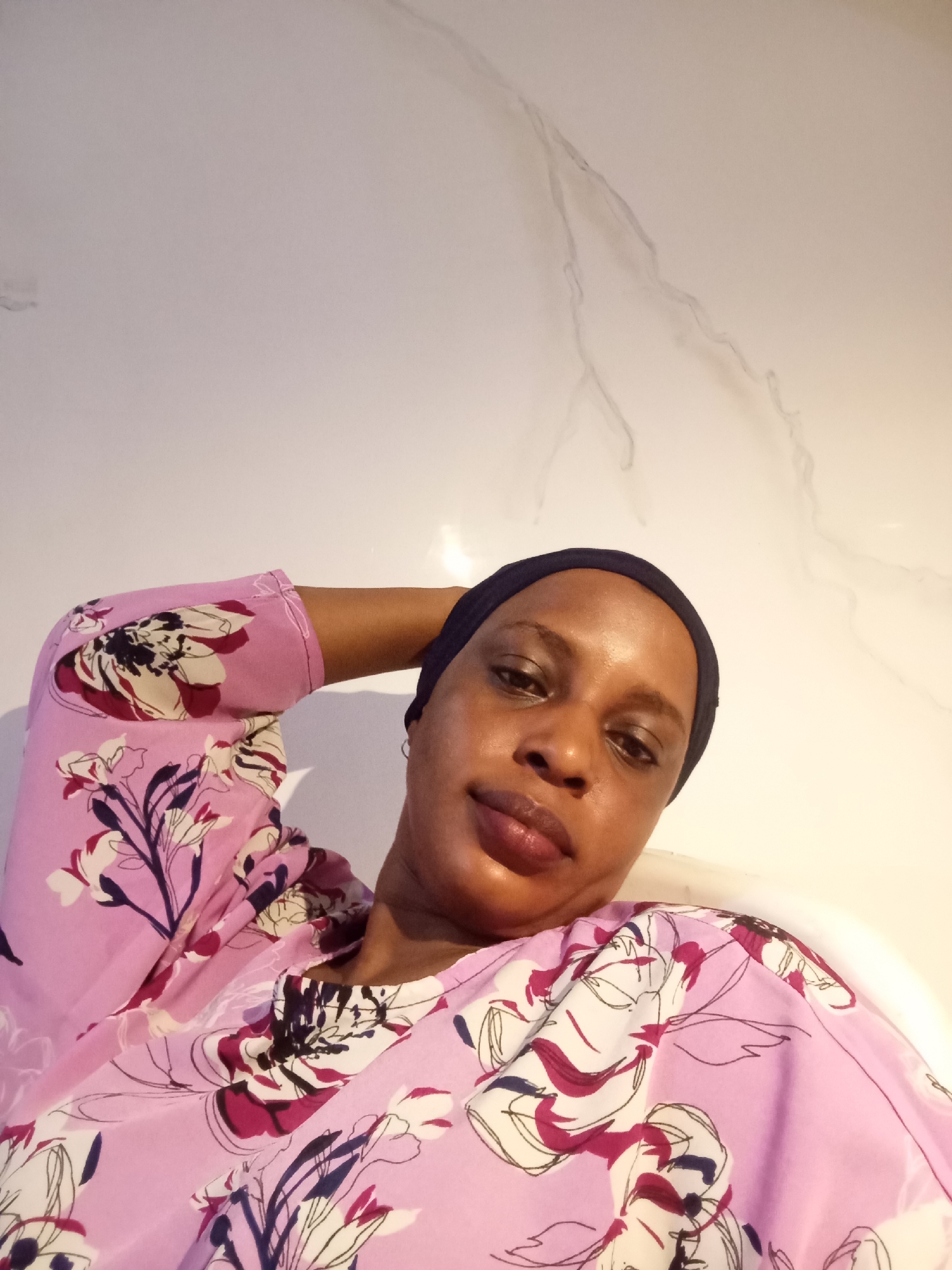 A medium shot captures a woman indoors, positioned against a white wall with marble-like veining. She wears a close-fitting, dark blue head covering and a pink, floral-patterned blouse. Her left arm is raised, the hand resting behind her head. The setting suggests a modern interior, possibly a bathroom or bedroom, although there is no additional context to confirm the specific location in Unguwar Rimi, Nigeria.