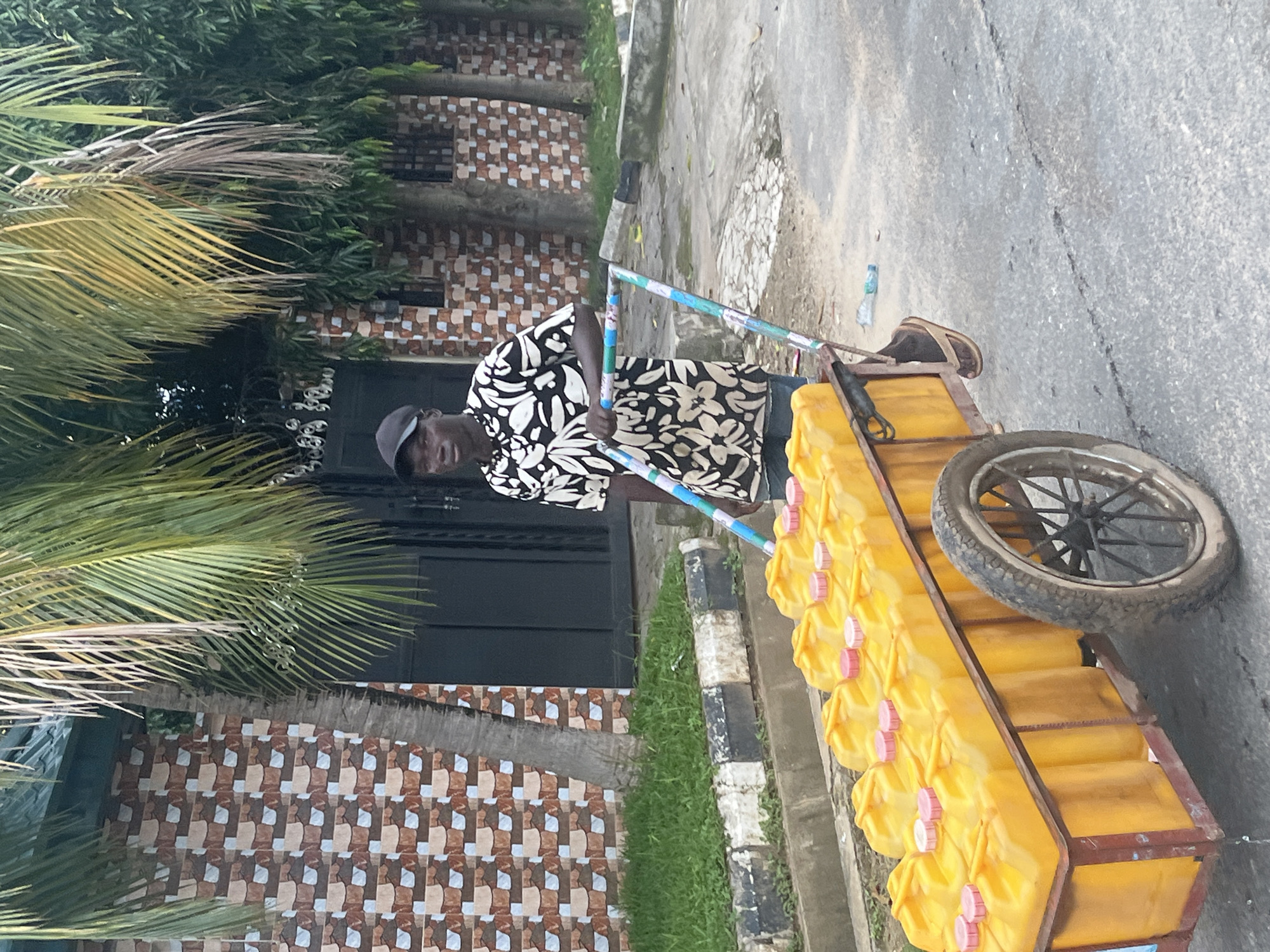 A young Black male wearing a black baseball cap and a short-sleeved black and white patterned shirt is pushing a cart laden with yellow jerrycans on a paved road. The cart has a metal frame with a single wheel at the back and appears to be made from a repurposed trolley. The jerrycans are packed tightly together, and some have red caps. The man is wearing brown sandals. The background shows a wall with a patterned facade of brown, beige, and white tiles, and some lush green vegetation including palm trees. The scene appears to be in an urban area, possibly a residential street, in Abuja, Nigeria, given the context provided. The overall impression is of a street vendor or delivery person engaged in their daily work.