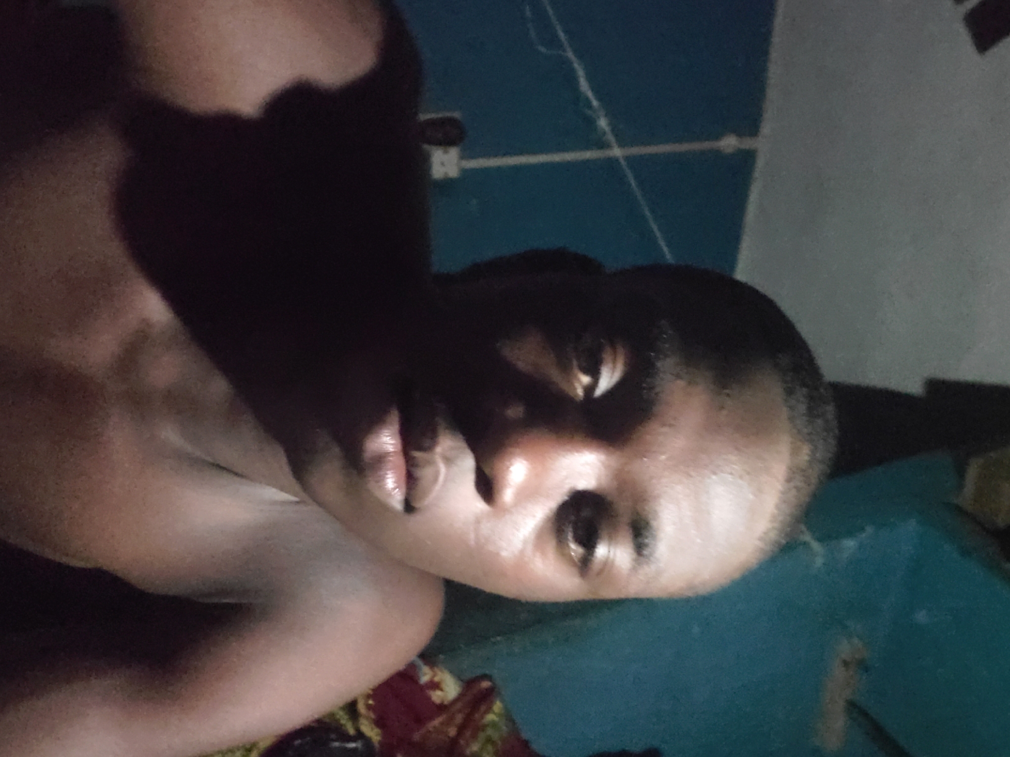 A dimly lit, close-up portrait of a person with dark skin is presented. The individual appears to be shirtless, with their face turned slightly towards the camera. The background includes portions of a wall with what seems to be electrical wiring or string visible. Parts of a bed or furniture with some fabric are also observable. The location is not identifiable as Unknown, Unknown.