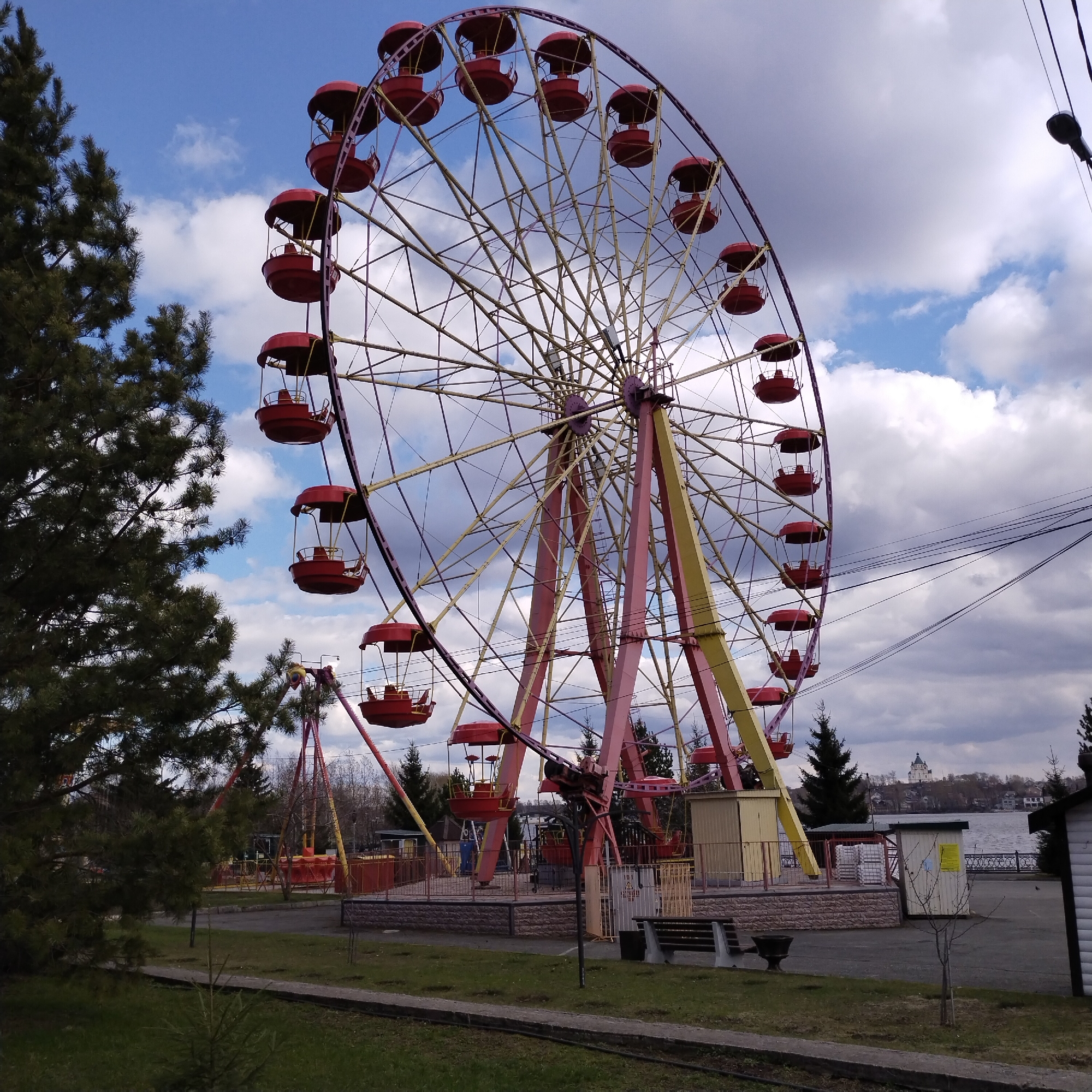 A clear day shows a Ferris wheel in Gorky Park, Nizhny Tagil, Russia. The Ferris wheel is large, with red enclosed cars and a yellow and pink structure. The wheel is positioned in the center of the frame, with a tall green pine tree to the left and the riverbank on the right. In the background, across the body of water, a white cathedral is visible on the skyline. A playground swing set is visible near the base of the Ferris wheel. In the foreground, a small section of grass lawn and a concrete path are visible. The sky is blue with scattered white clouds. A wooden building and telephone wires are near the right side of the image.